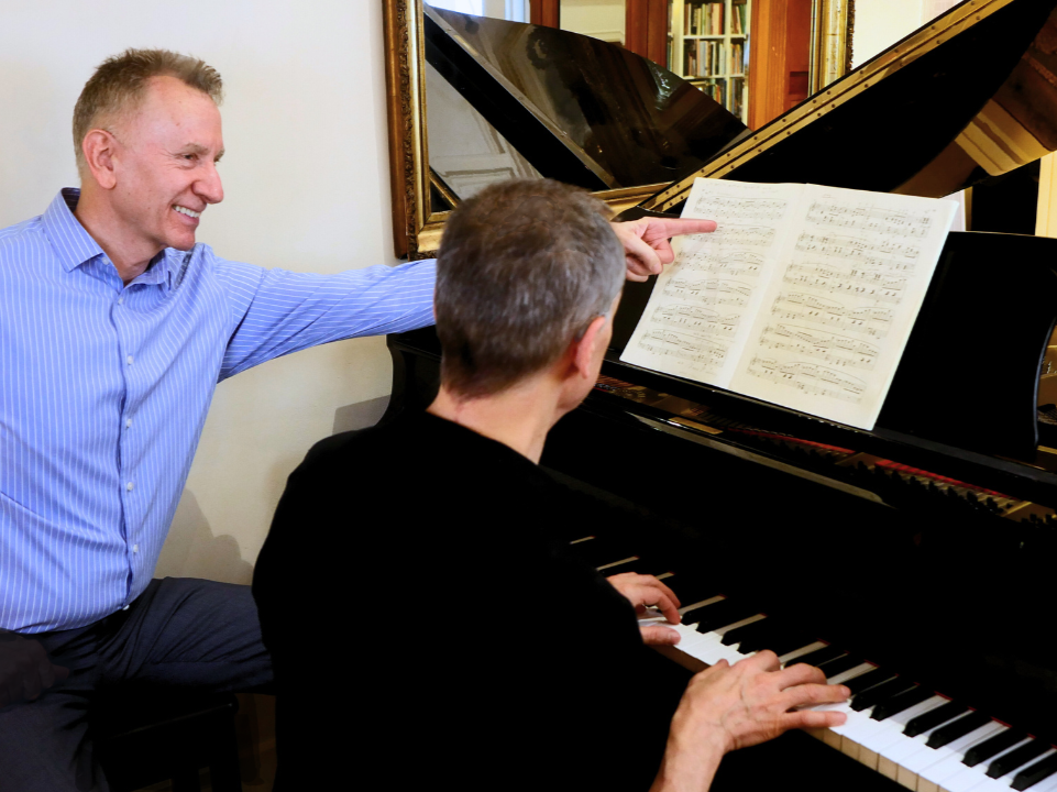 Two men at a piano, one playing and the other pointing at sheet music, in a room with a bookshelf and a mirror.