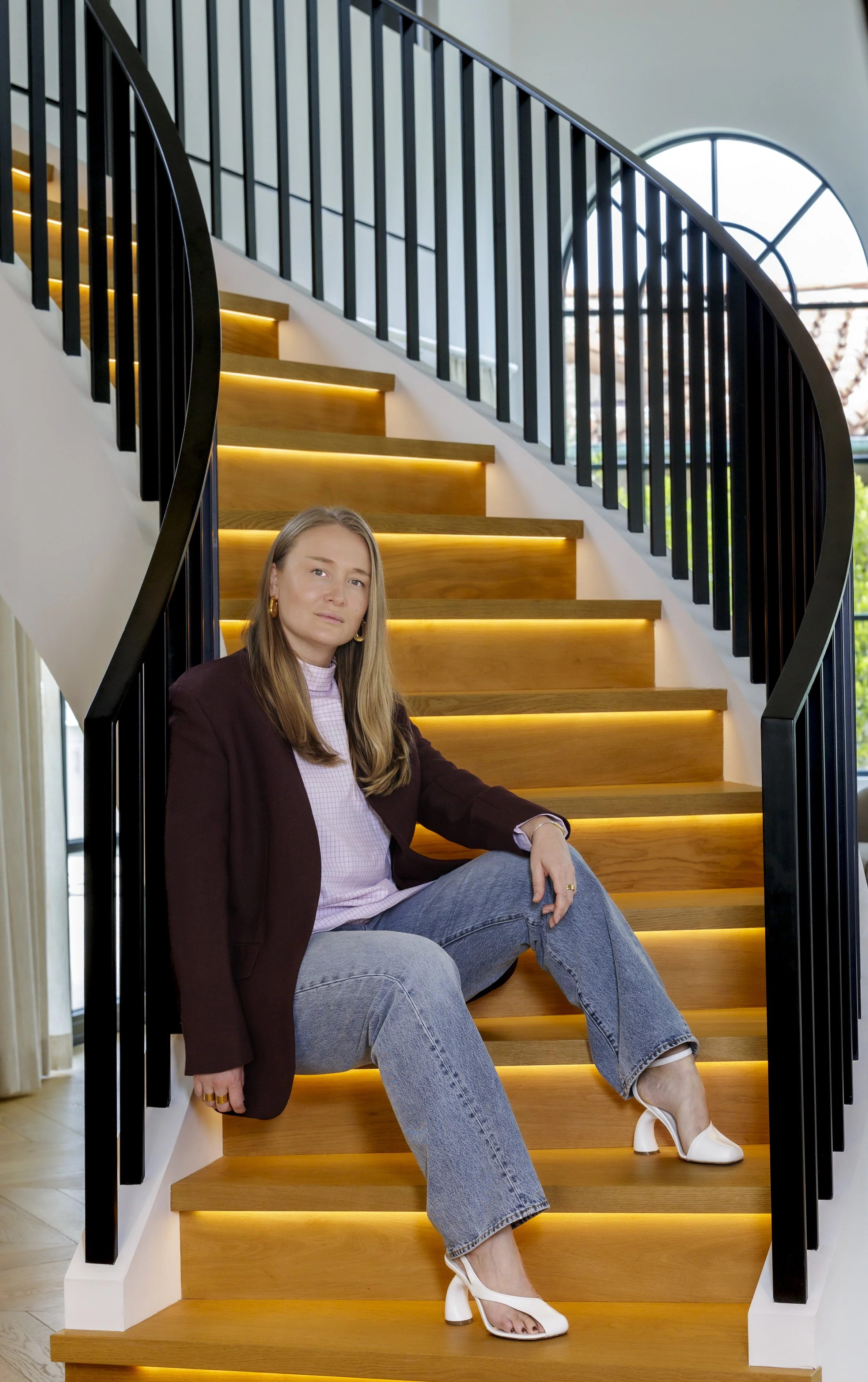 A woman sitting on a staircase with wooden steps and black railing, illuminated by yellow lighting, inside a modern home with large arched window in the background.
