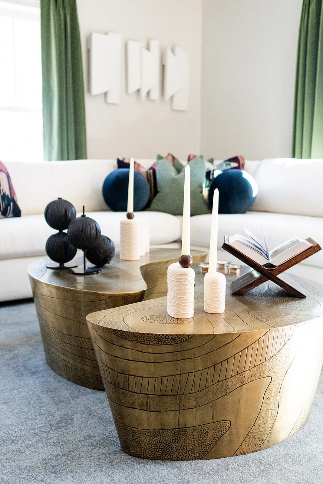 A contemporary living room featuring a white sofa, decorative brass nesting tables, assorted pillows, green velvet drapes, and sculptural art on the wall.