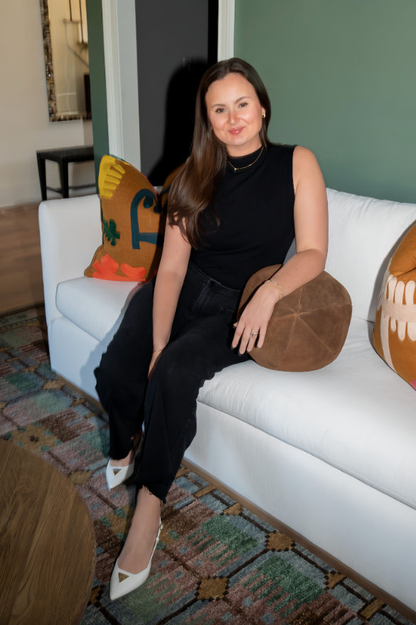 Smiling woman with long brown hair wearing a black shirt and gold jewelry indoors.