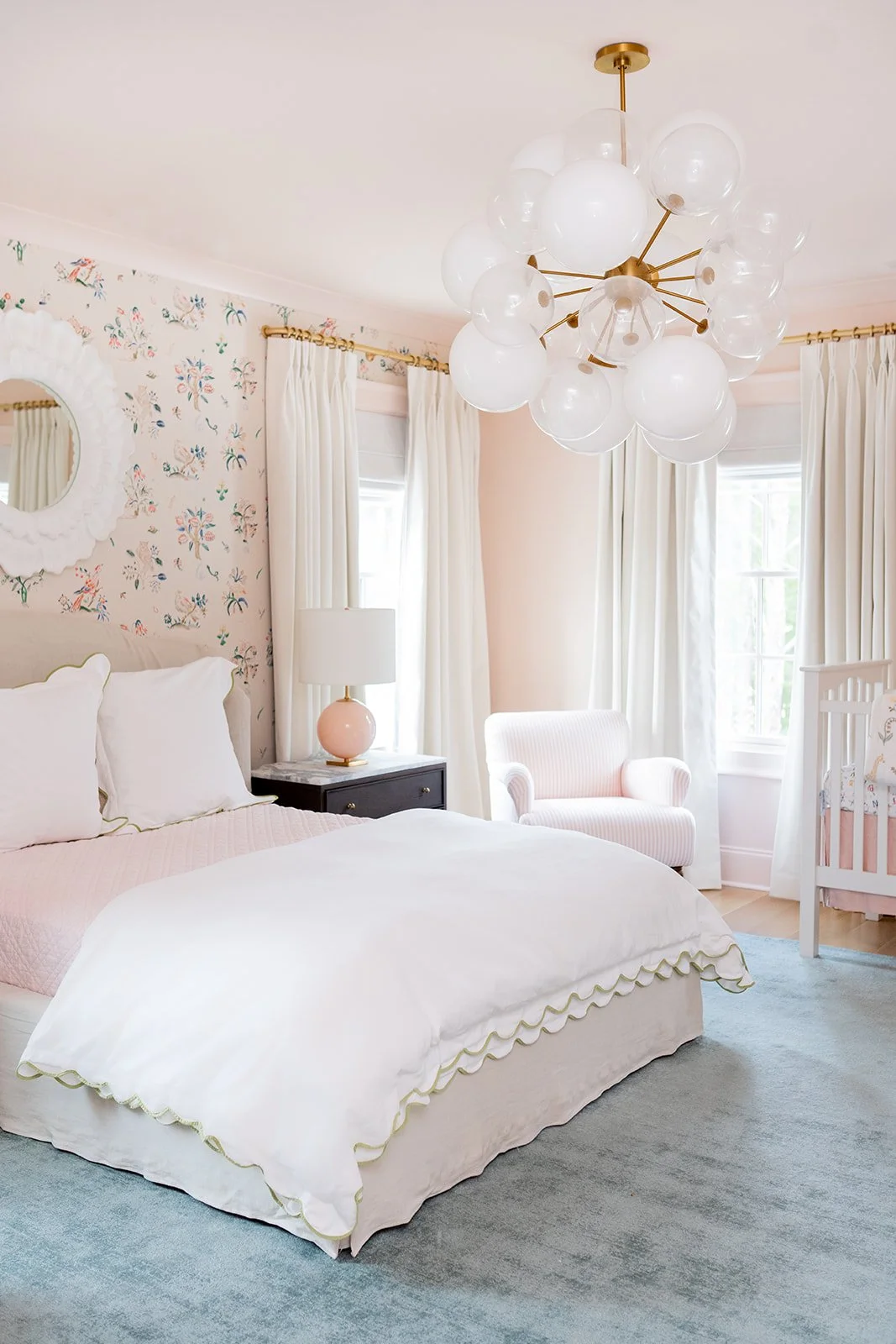 A bedroom with a white bed, pink bedding, and a scalloped quilt, a black nightstand with a pink lamp, a white armchair, white curtains, floral wallpaper, and a modern chandelier with glass globes.