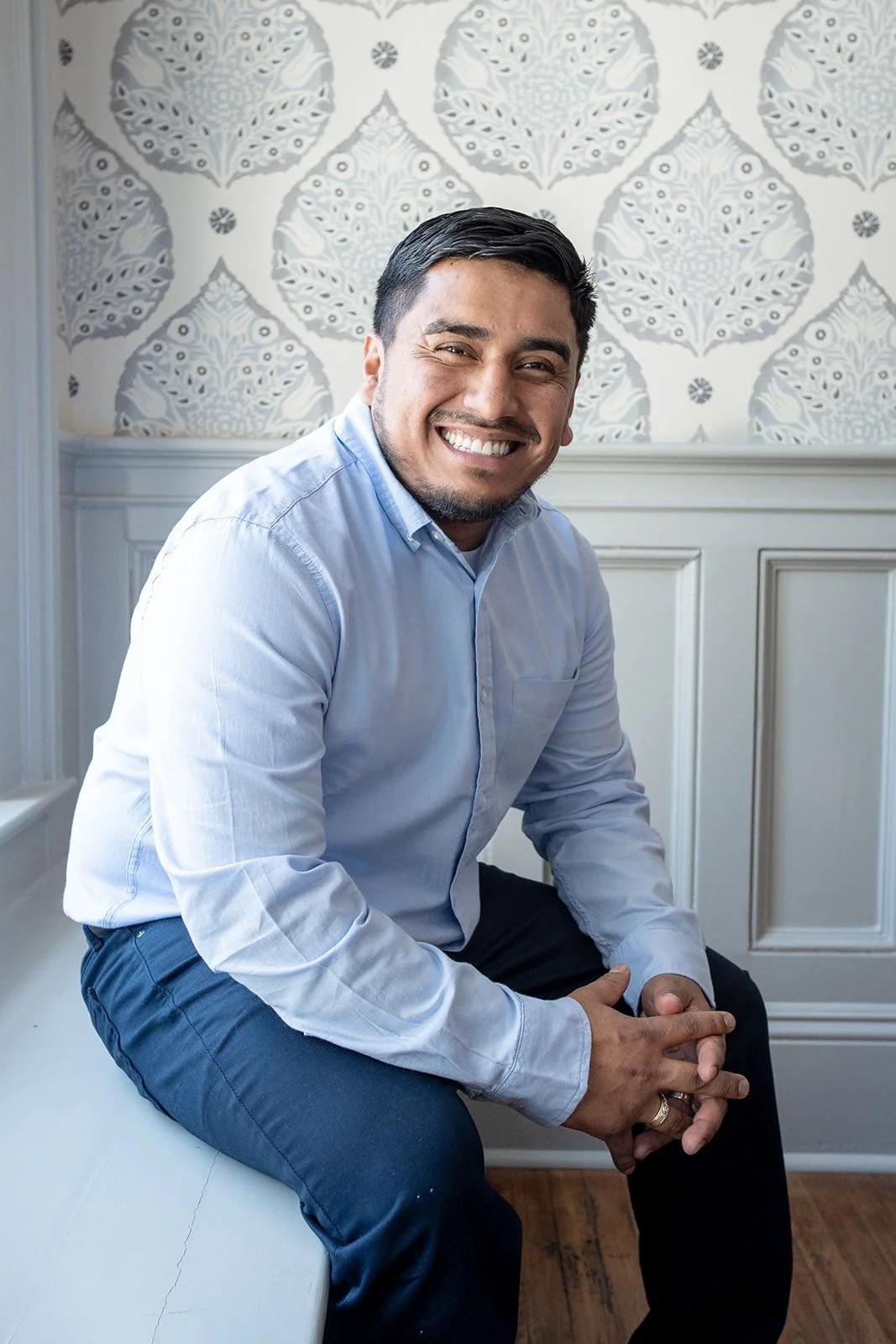 A hispanic man in a blue button down shirt smiles in front of a wallpapered wall