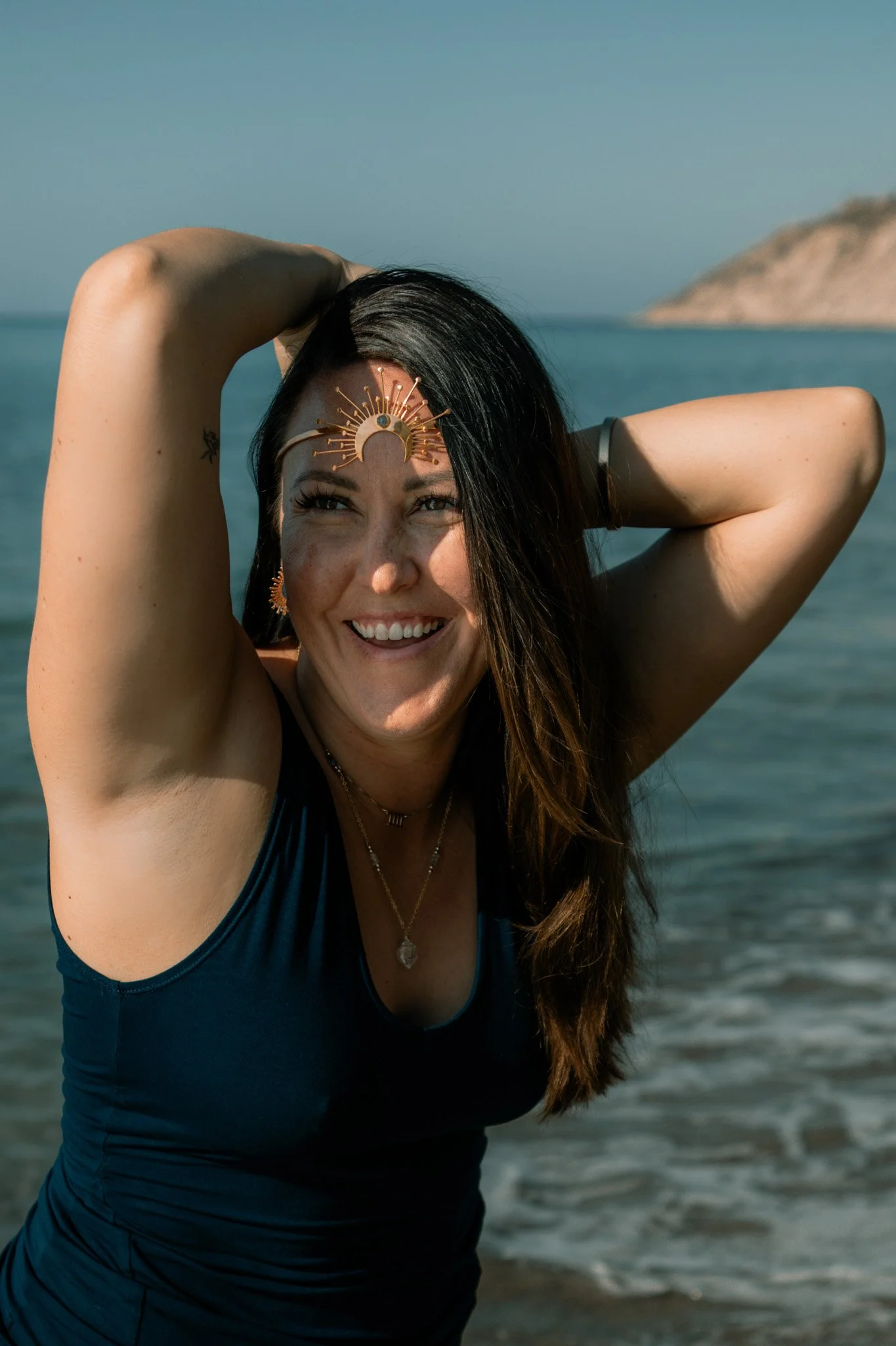 A woman smiling outdoors with sunlight on her face, wearing a black top and layered necklaces.