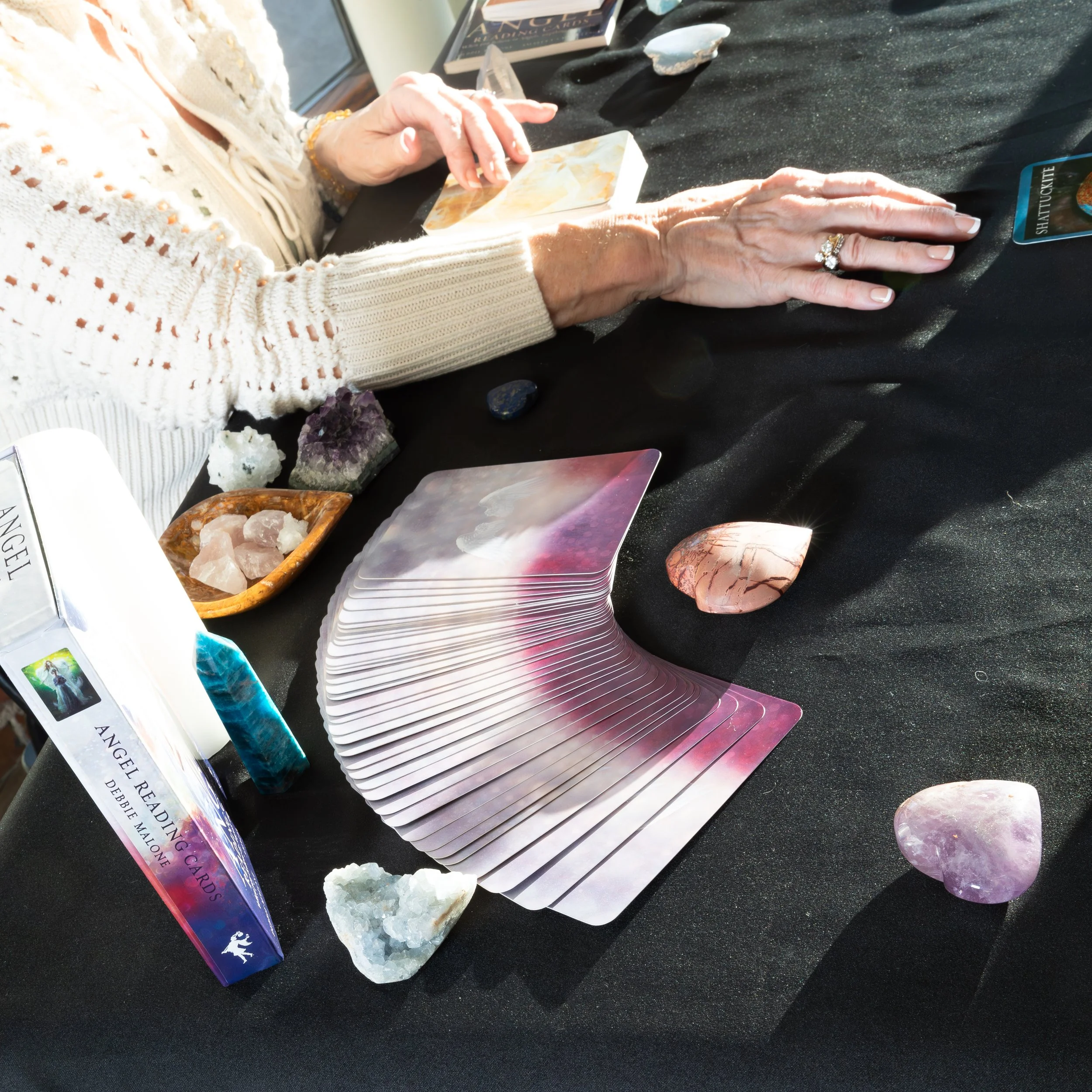 A person at a table displaying a tarot card deck, crystals, and stones, with their hand on a tarot card and the other hand resting on the table, wearing rings and a bracelet, in a well-lit room.
