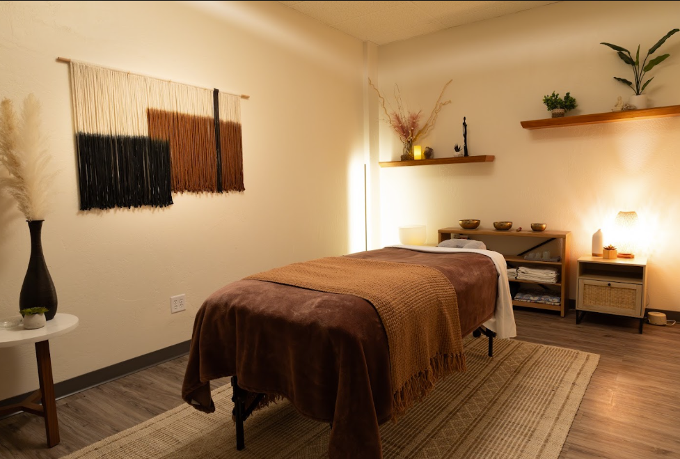 A massage room with a massage table covered in brown blankets, surrounded by warm lighting, shelves with decorative items, and plants.