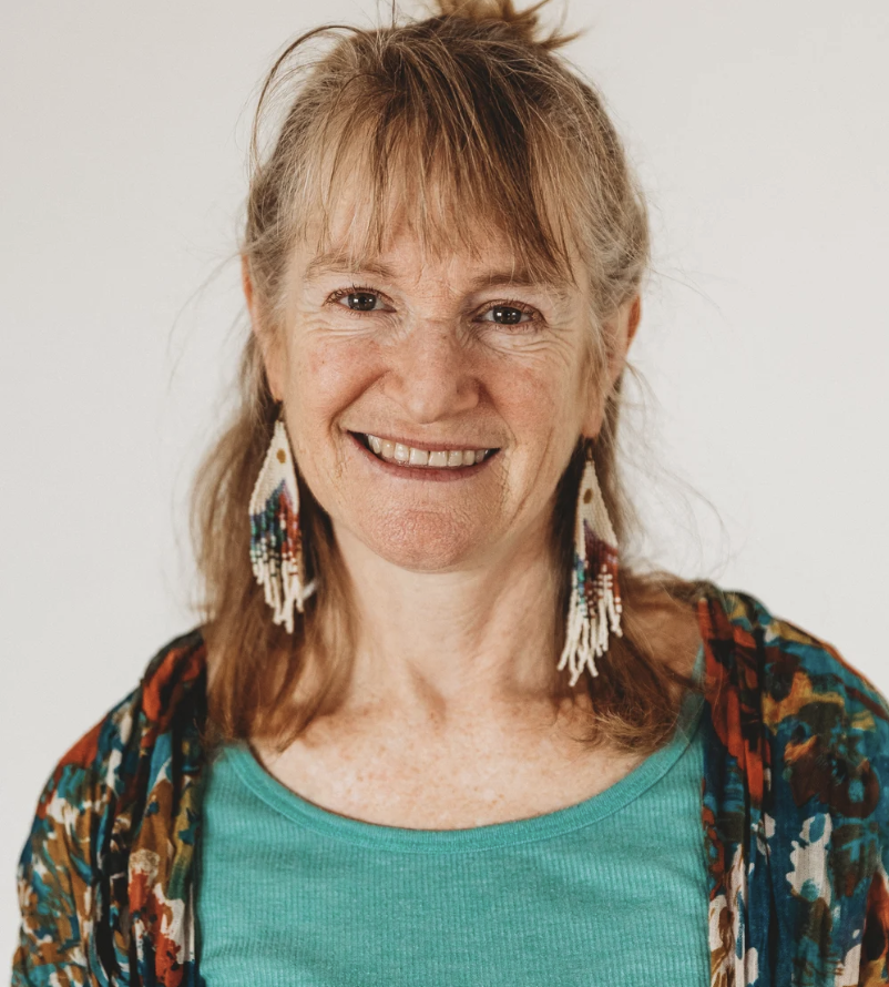 A smiling woman with shoulder-length red hair, wearing beaded earrings and a patterned jacket over a teal top, against a plain white background.