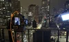 Person taking a photo with a camera on a balcony at night, overlooking a city with tall buildings and illuminated windows.