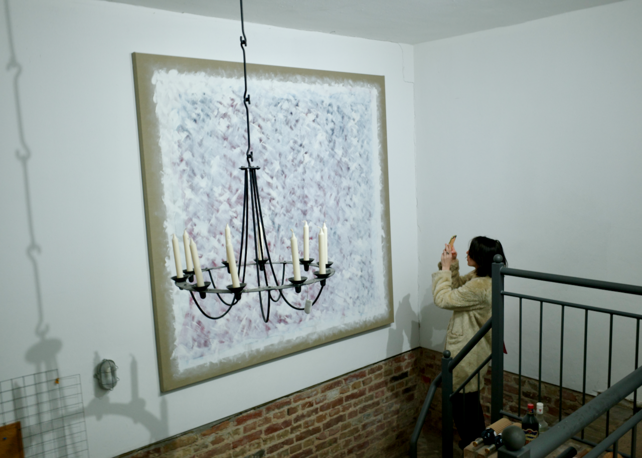 A woman taking a photo or video with a smartphone inside an art gallery or studio. The room has white walls, a large abstract painting with white and pink brushstrokes, an iron chandelier with candles, a brick wall detail, and some items on the floor