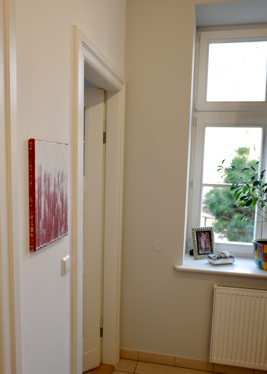 A corner of a room with a white door frame, a artist canvas on the wall, a window with a plant and picture frame on the windowsill, and a radiator below the window.