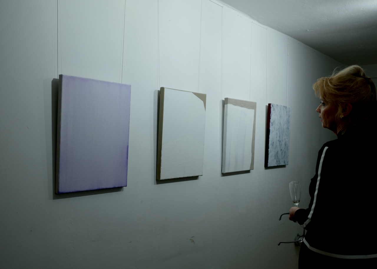A woman looking at a series of abstract paintings on a white gallery wall, holding a glass of champagne.
