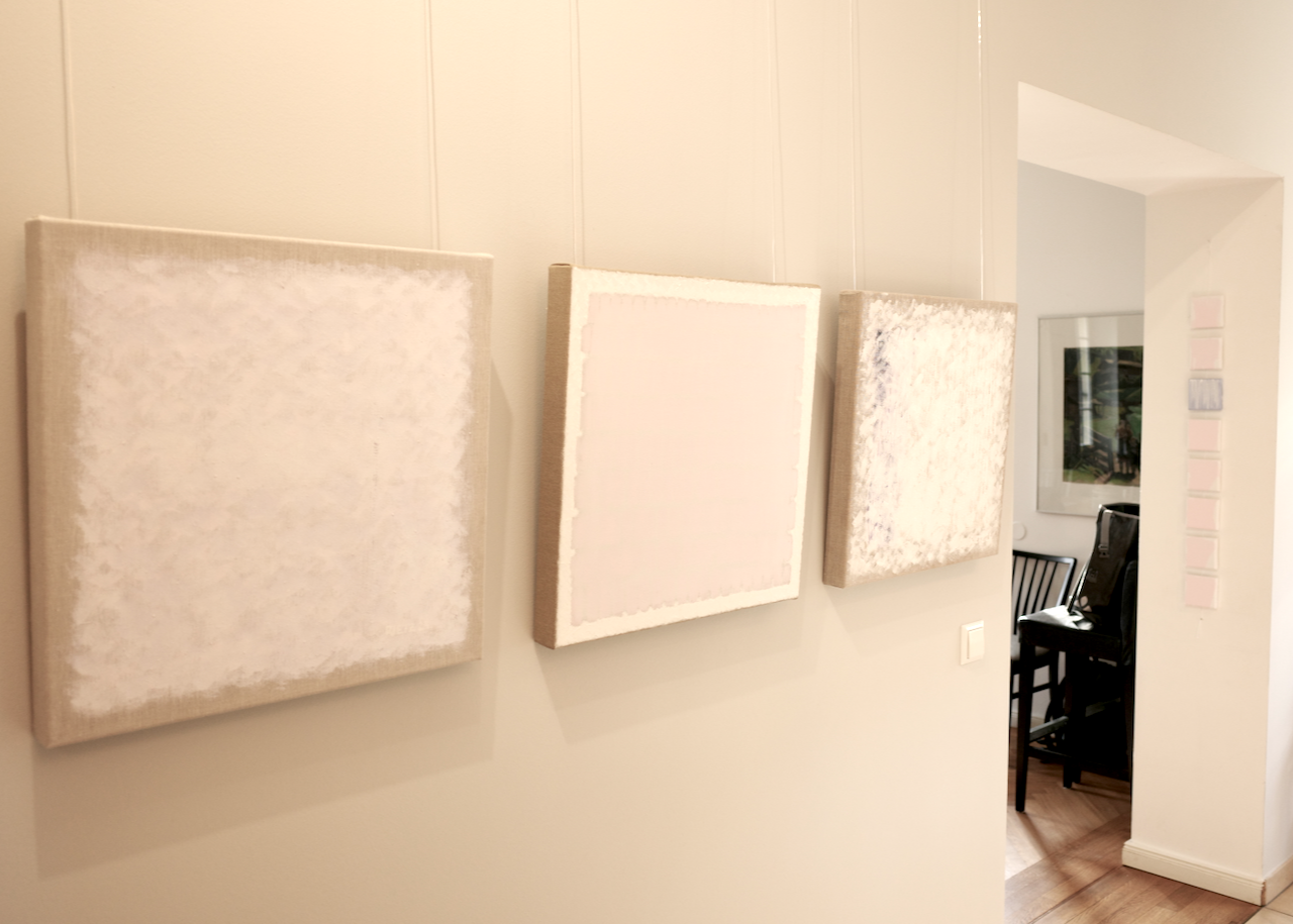 Three blank canvas paintings hung on a light-colored wall in a room, with a doorway leading to a dining area with black chairs and a window.
