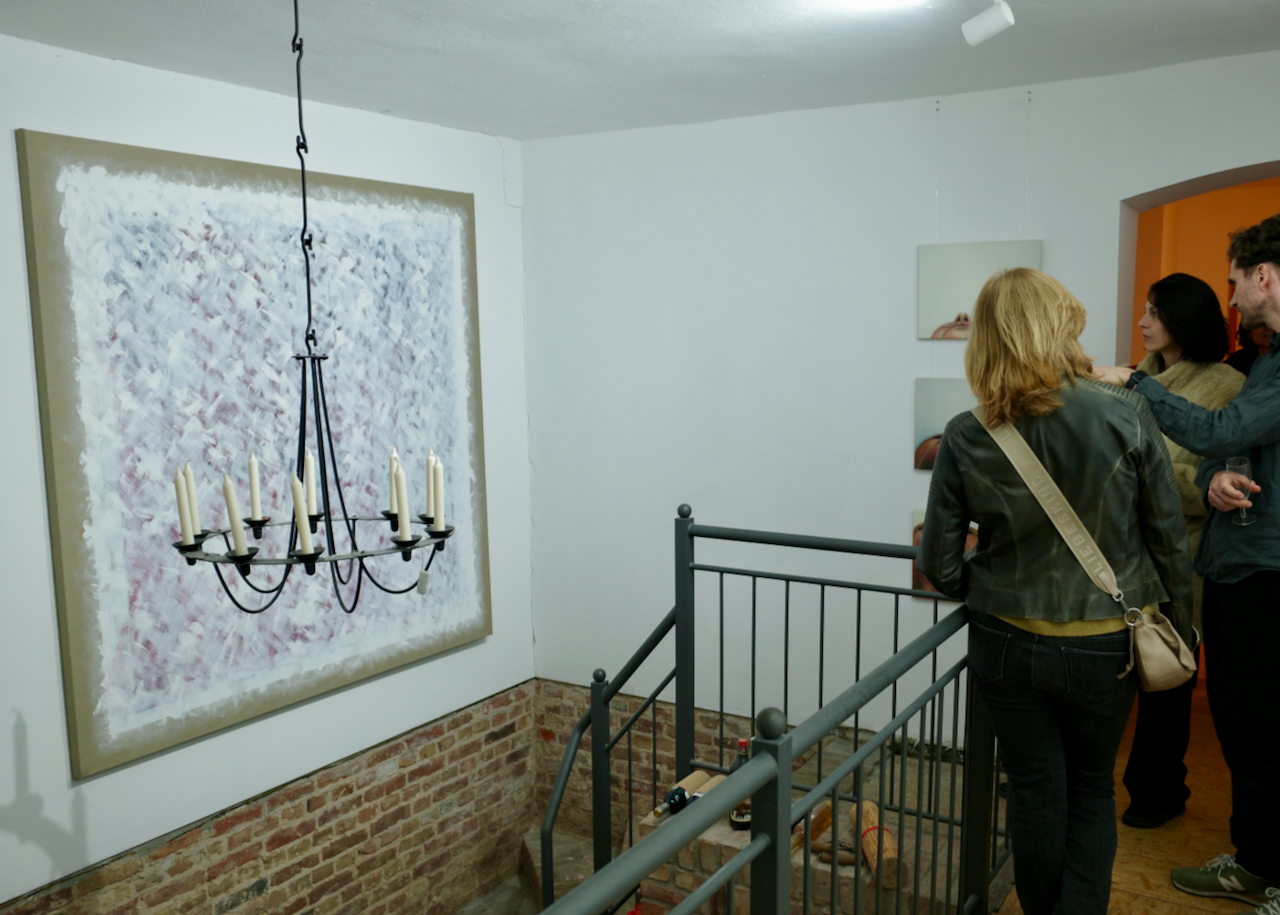 People viewing abstract artwork at an art gallery, with a chandelier hanging from the ceiling and a brick wall at the bottom of the white wall.