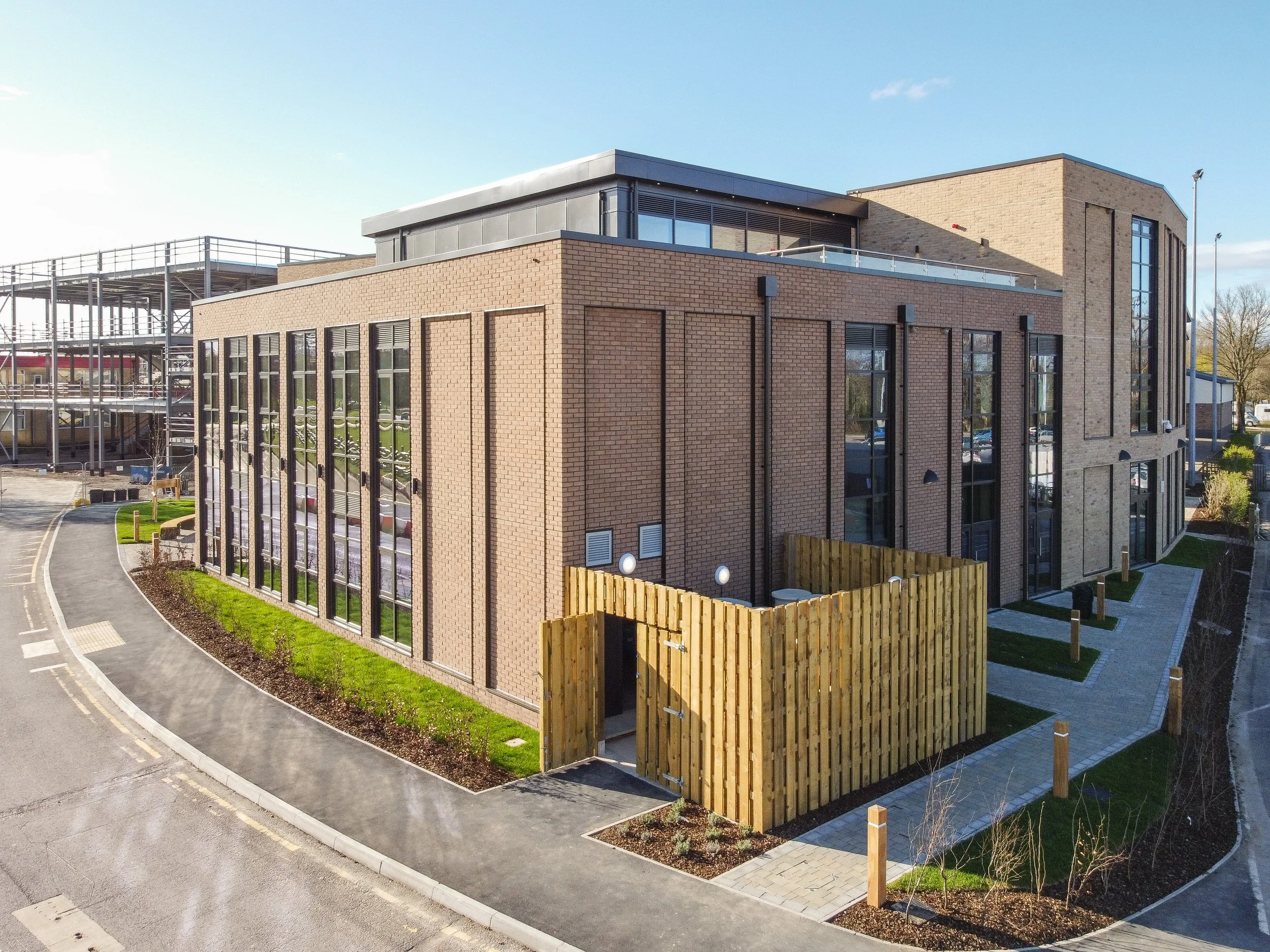 A modern multi-story building with brick and large glass windows, surrounded by a paved walkway, small green areas, and a wooden fence section.