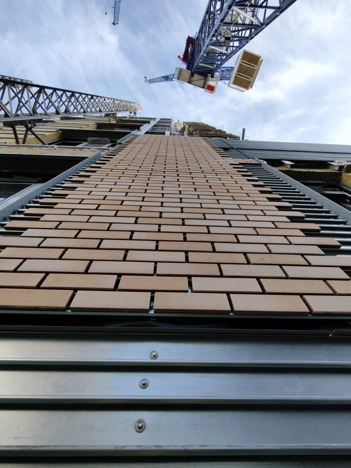 Low-angle view of a tall building under construction with brick facade, construction crane at the top lifting a crate, and blue sky in the background.