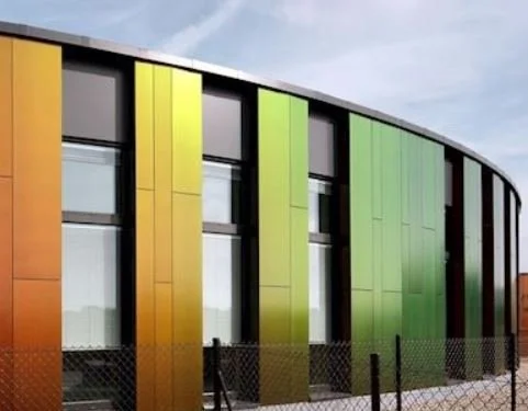 Modern building with multicolored vertical panels in shades of orange, yellow, green, and black, with large glass windows and a fenced perimeter.