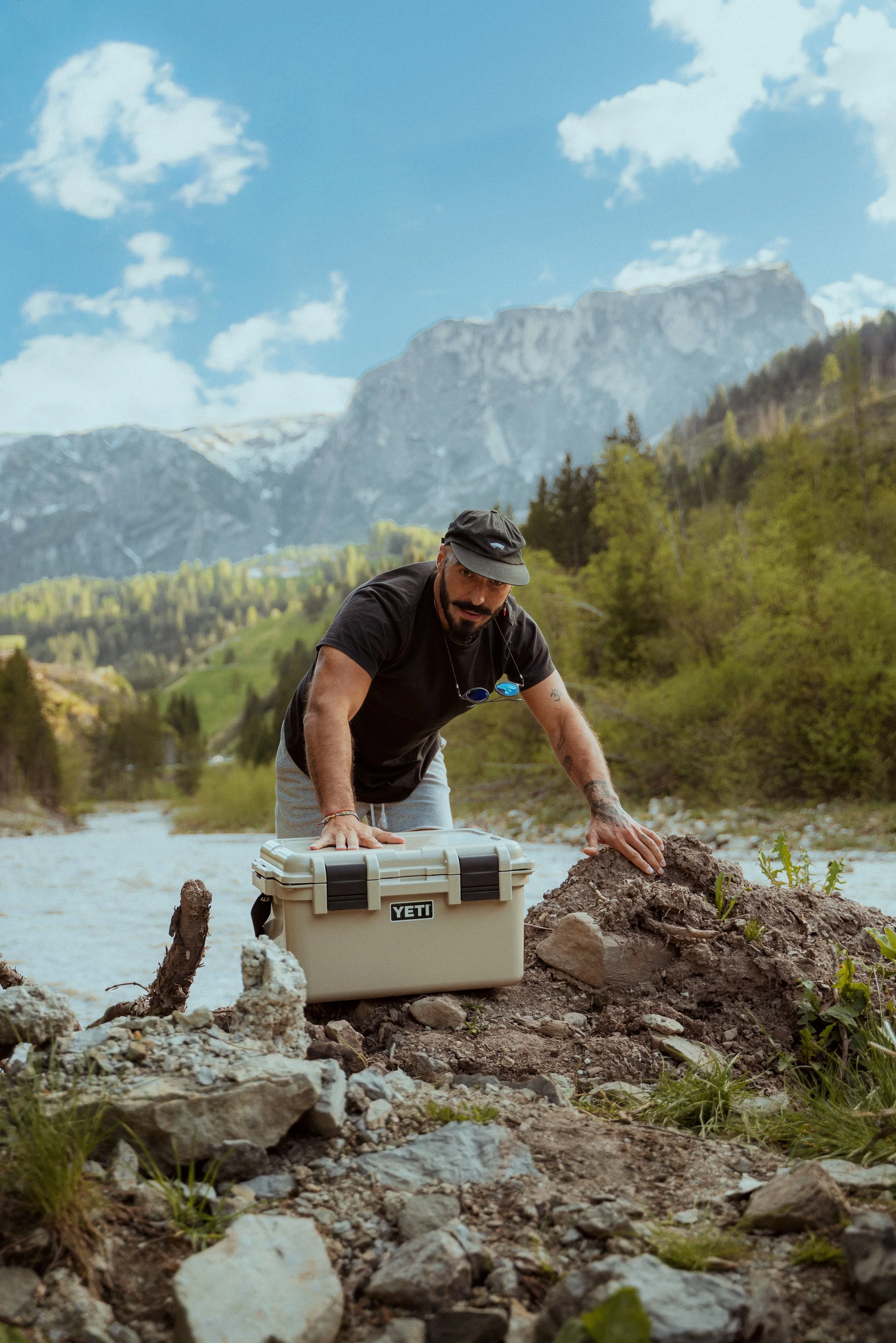 A man wearing a black shirt and hat kneels on the ground outdoors by a river, carrying a YETI storage box with greenery, with mountains and trees in the background.