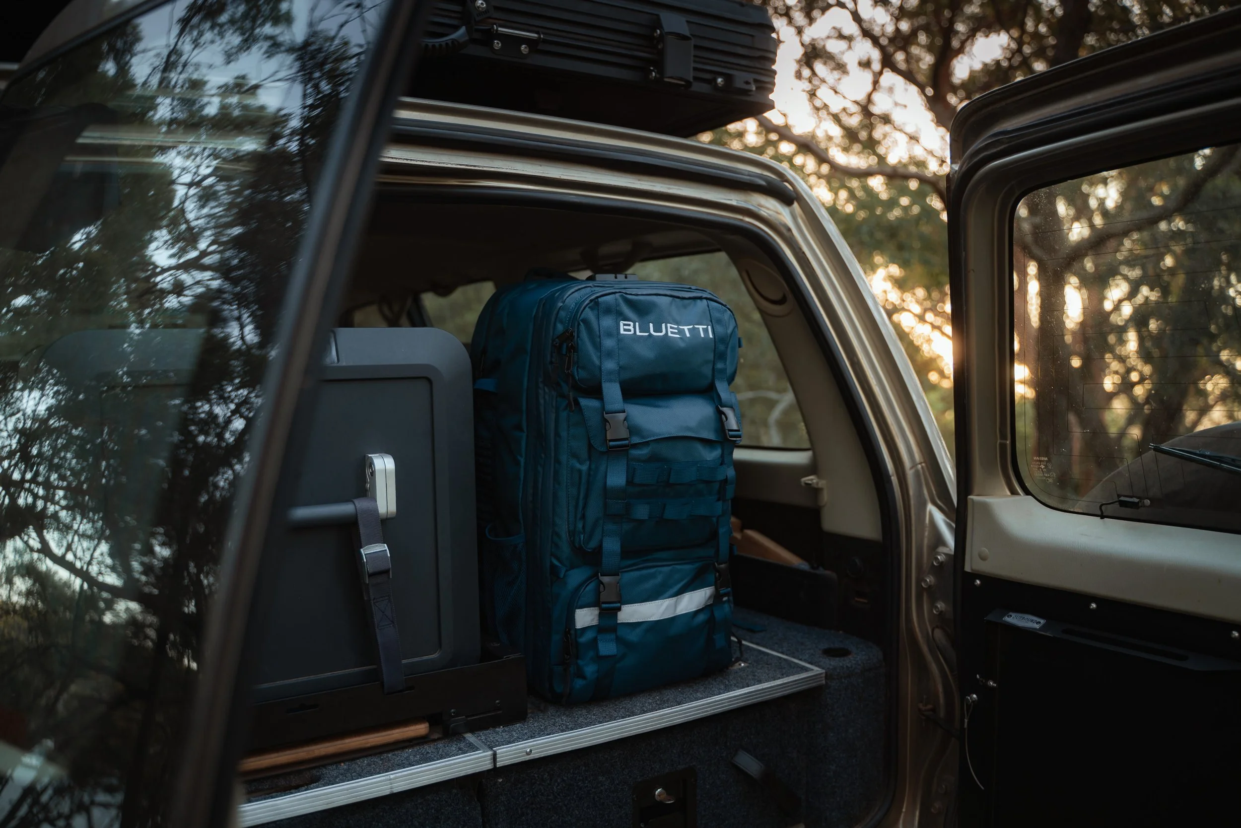 Open vehicle trunk with a blue backpack labeled 'BLUETTI' inside, surrounded by trees and a setting sun.