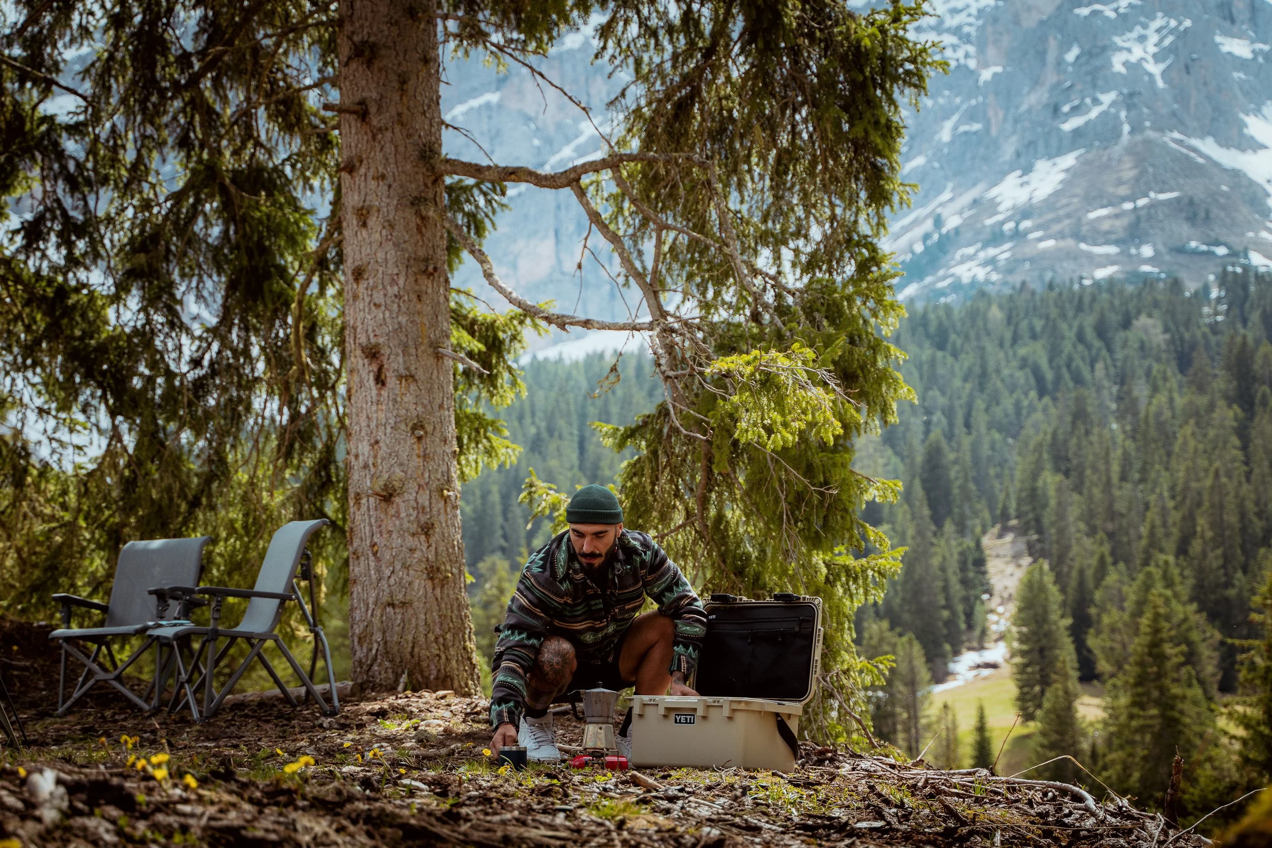 A man with a beard and tattoos, wearing a beanie and patterned jacket, is crouched down outdoors in a forested mountain area, preparing equipment from a YETI cooler with trees and mountains in the background.