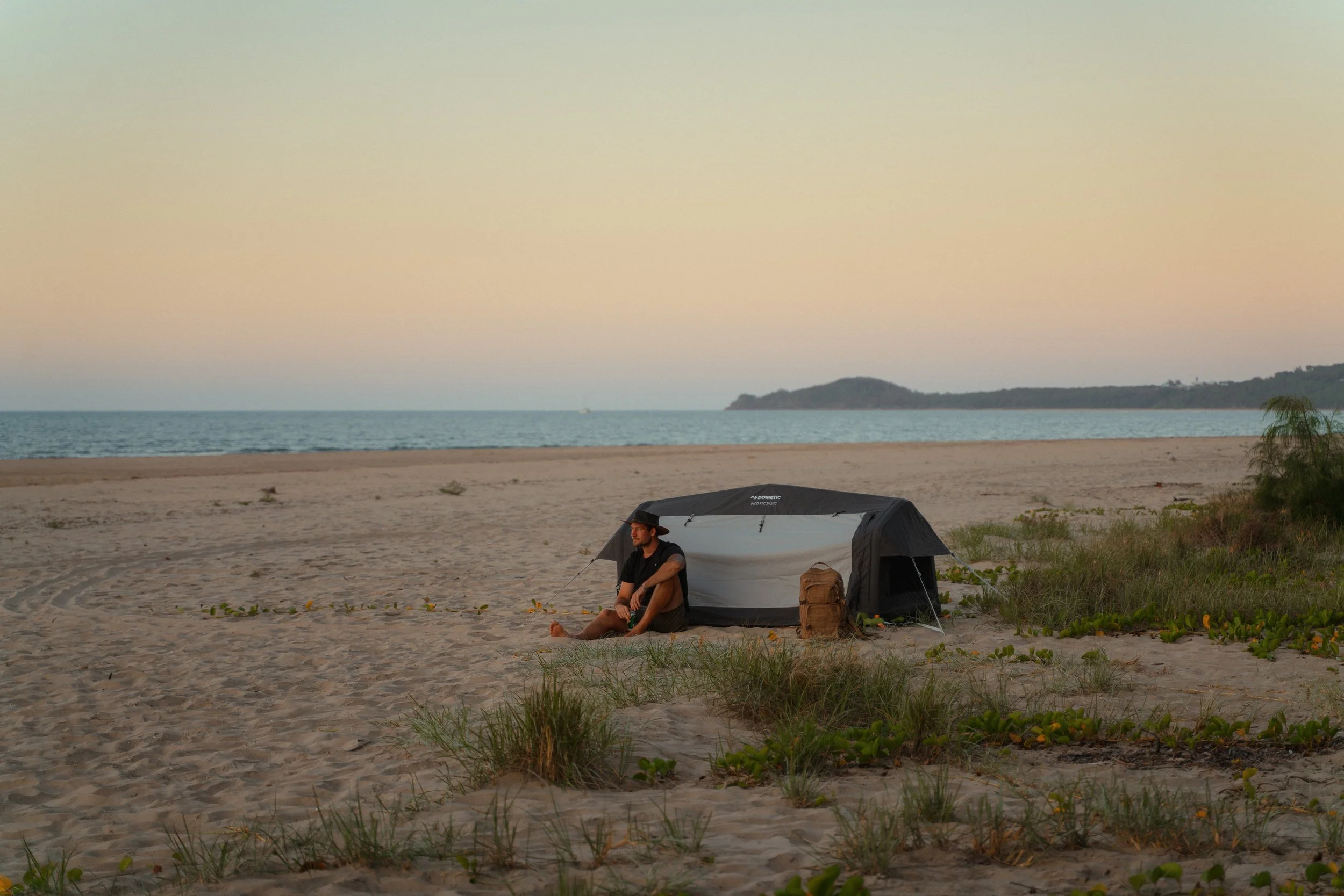 A person sitting outside a tent on a sandy beach at sunset, with the ocean and distant hills in the background.