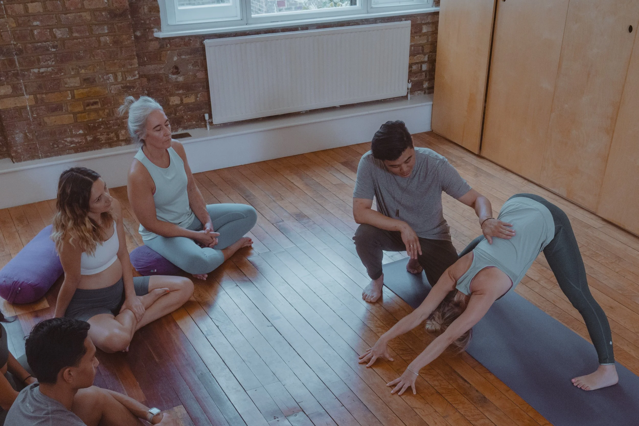A woman performs a downward dog yoga pose on a mat while a man adjusts her posture, surrounded by three women and a man sitting on the wooden floor watching her in a yoga studio.