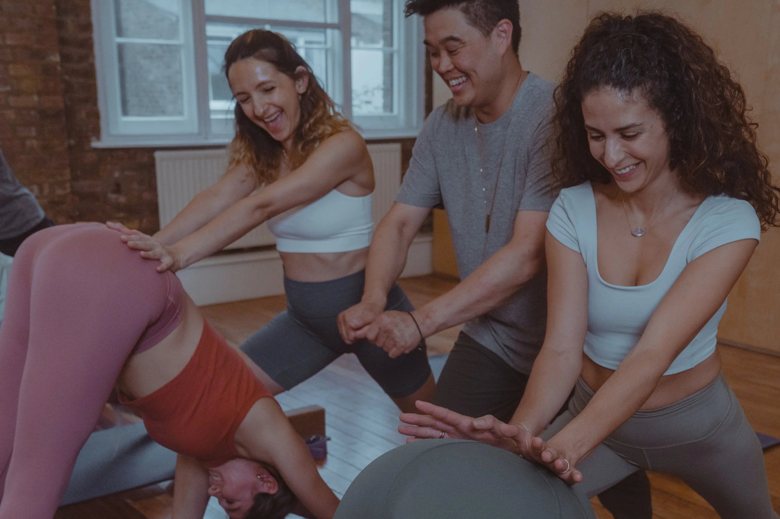 People participating in a yoga class, stretching and having fun in a studio with wooden floors and brick walls.