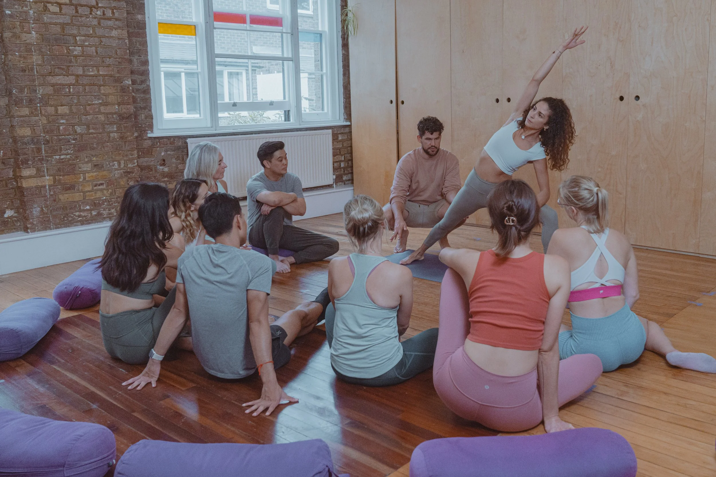 A group of people sitting on the wooden floor of a studio, attentively watching a woman demonstrate a yoga pose while lying on one side with one arm extended overhead.