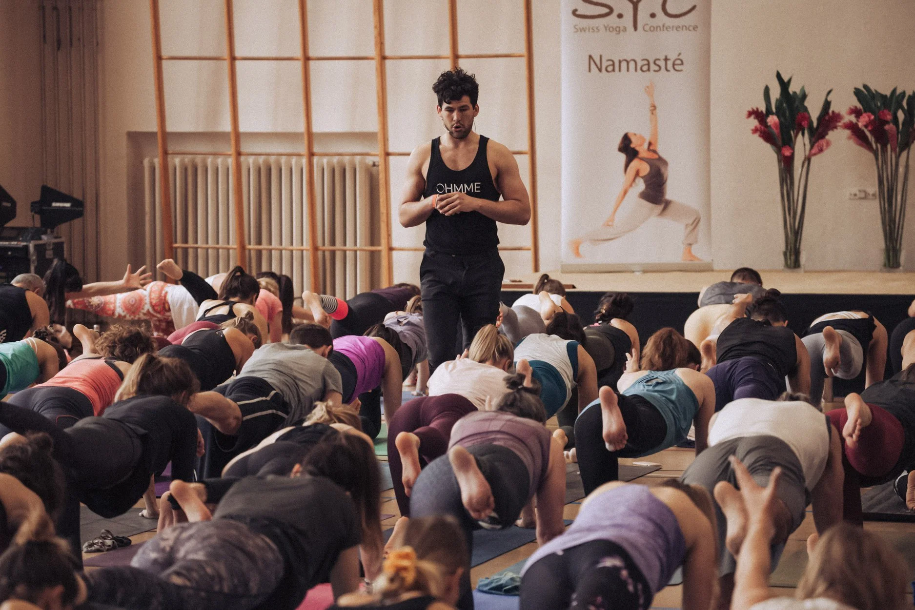 A group of people participating in a yoga class in a studio, led by an instructor standing at the front, with a backdrop featuring a banner with the word "Namaste" and a woman in a yoga pose.