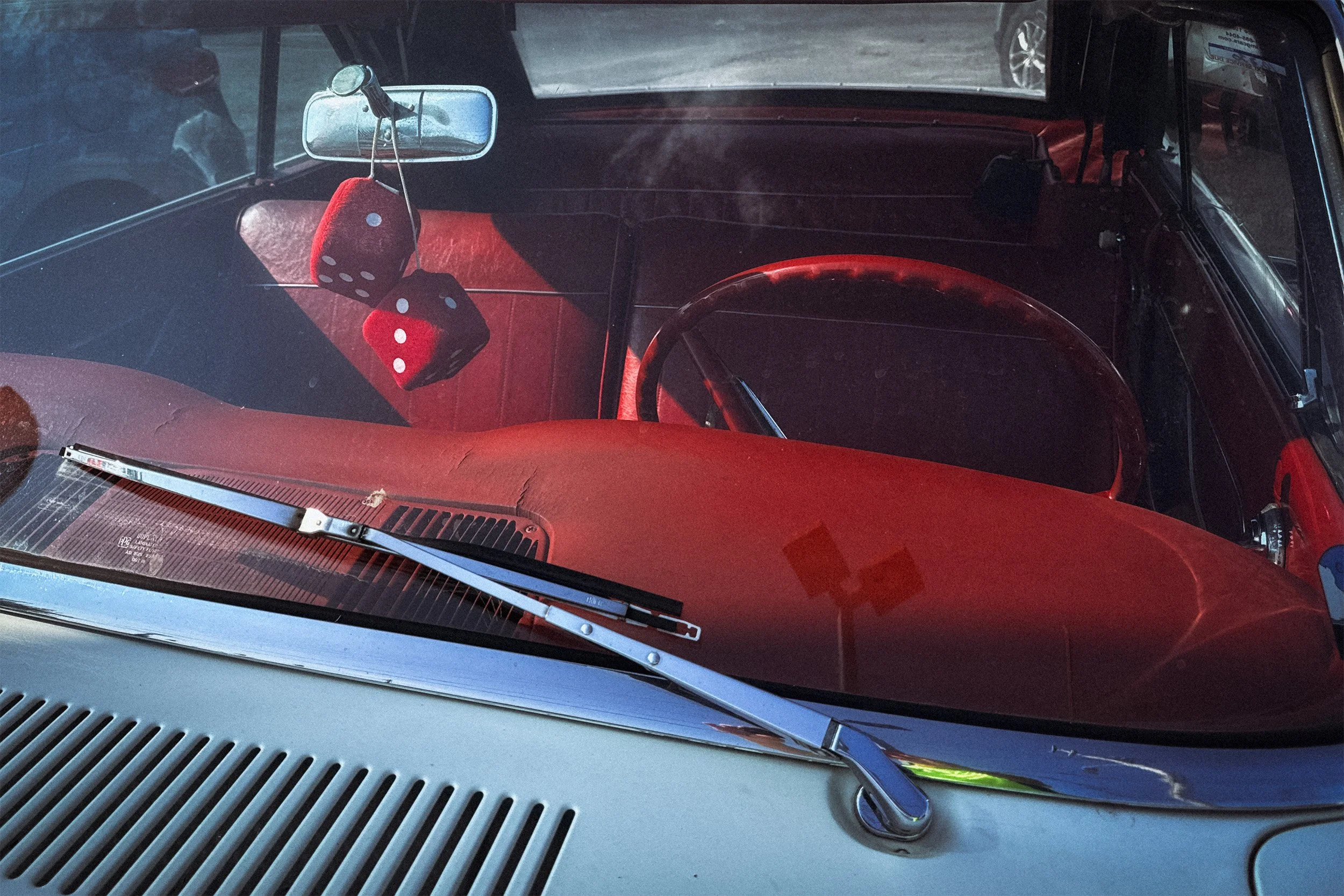 Image of the red interior of a 1960's car taken through the windshield with red fuzzy dice hanging from the rear view mirror