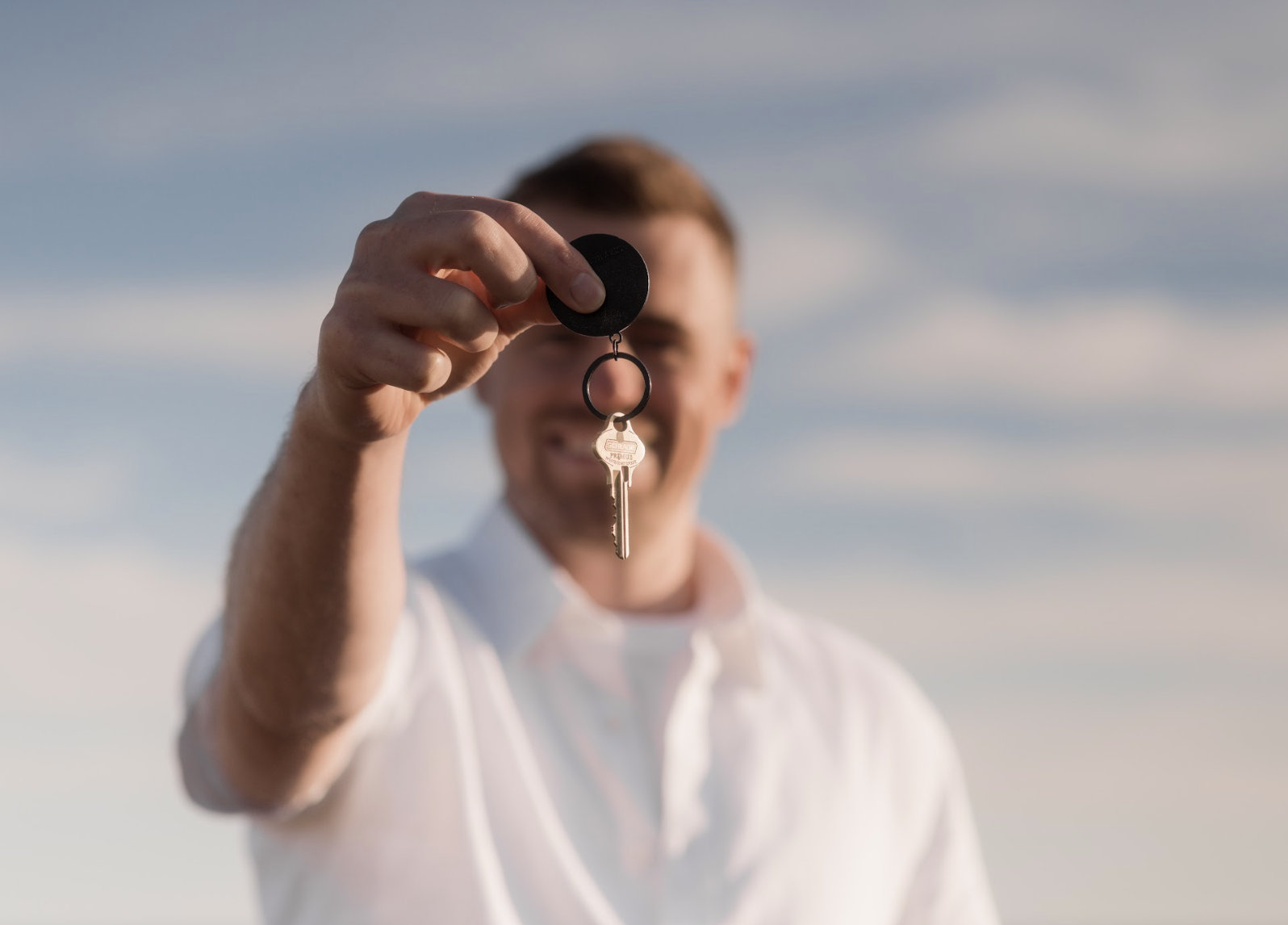 Smiling man in white shirt holding a key with a black keychain against a partly cloudy sky.