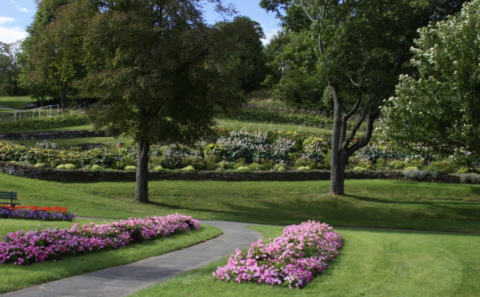 A peaceful park scene with a paved walking path, colorful flower beds, lush green grass, and tall trees under a blue sky.