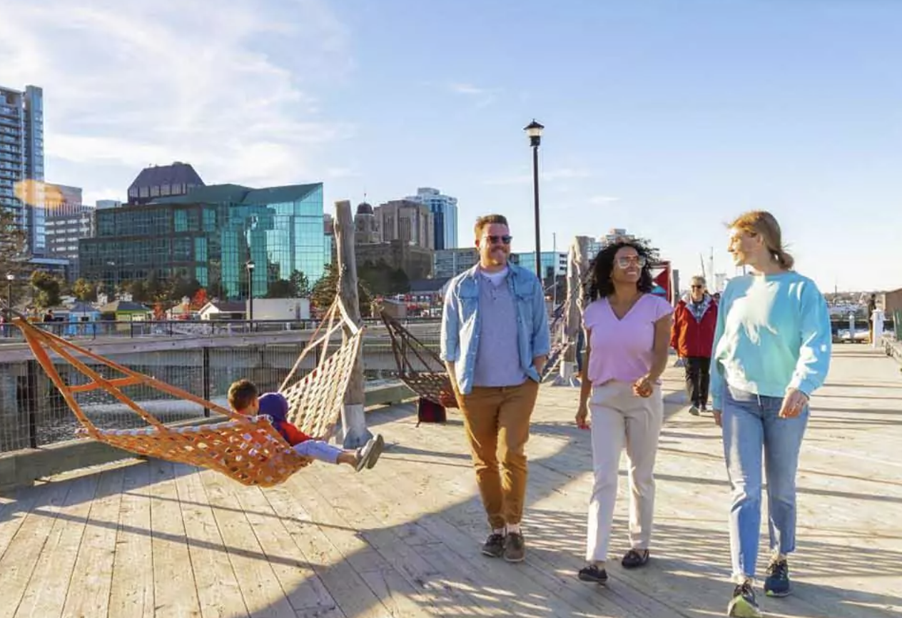 Three people walking on a Halifax boardwalk near with city buildings in the background, while a child lies on an orange hammock.