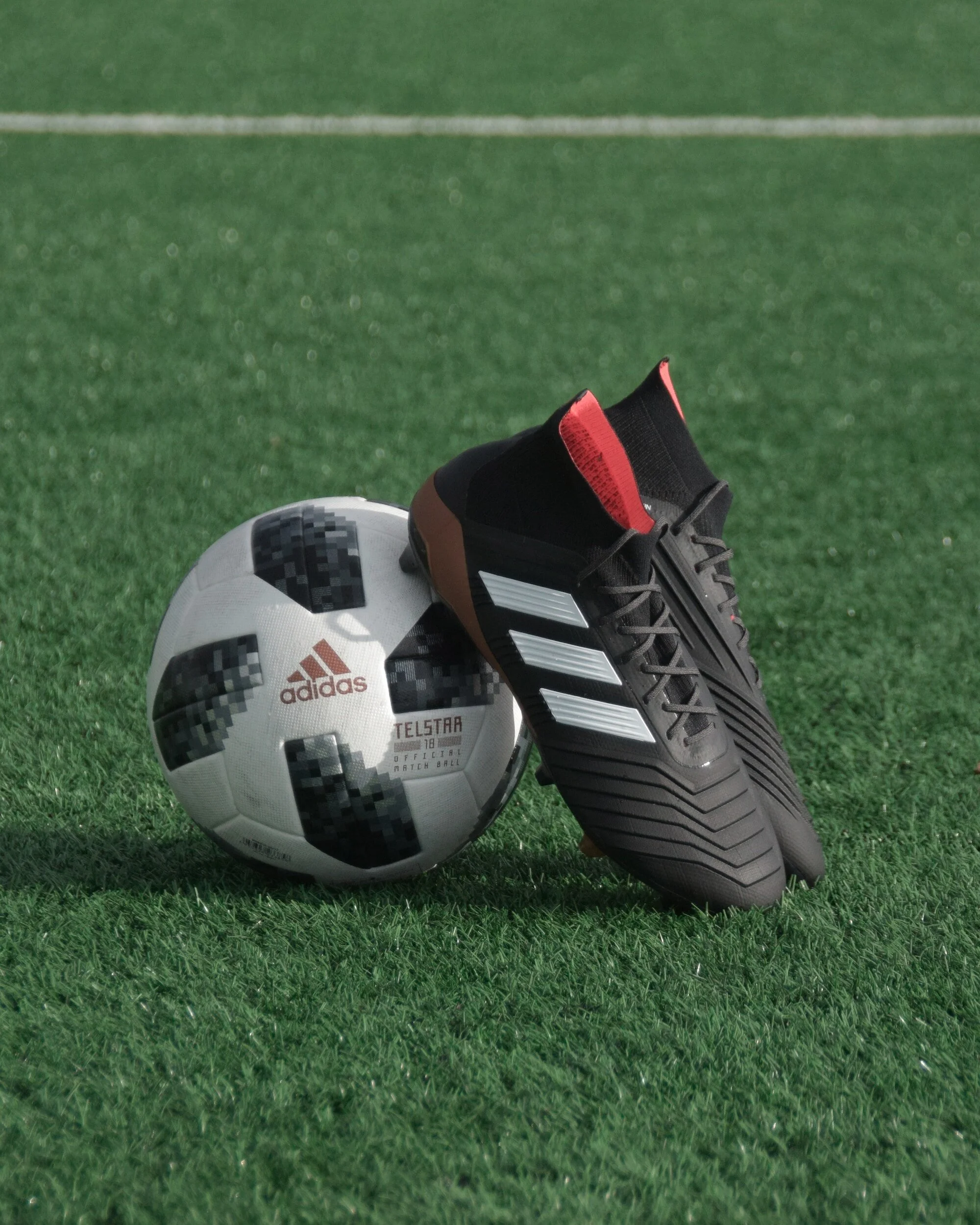 Ballon de football Adidas à côté d'une paire de chaussures de sport noires avec des bandes blanches, posés sur un terrain en herbe.