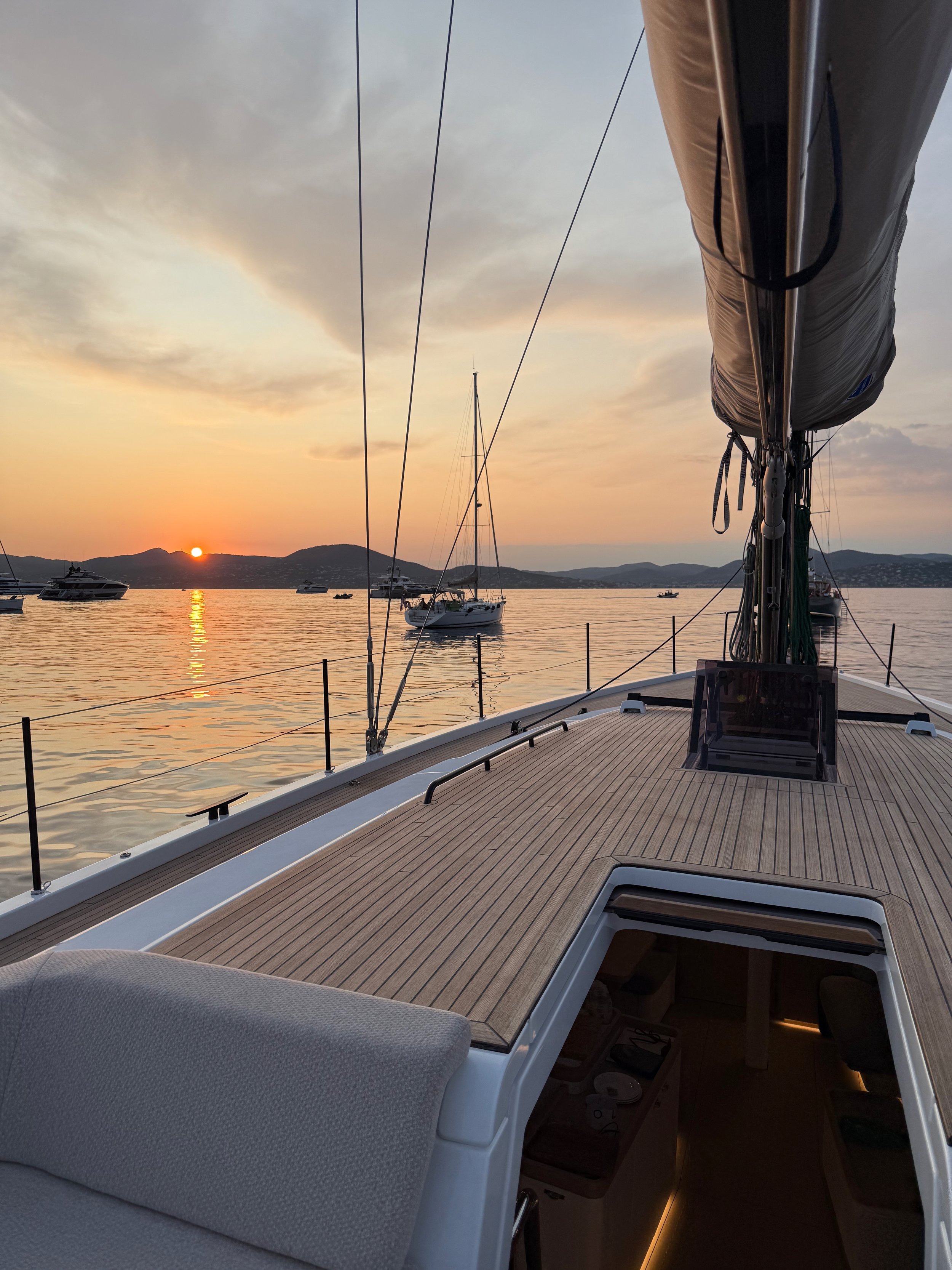 Sailboat deck overlooking anchored yachts and calm water at sunset, with mountains in the background