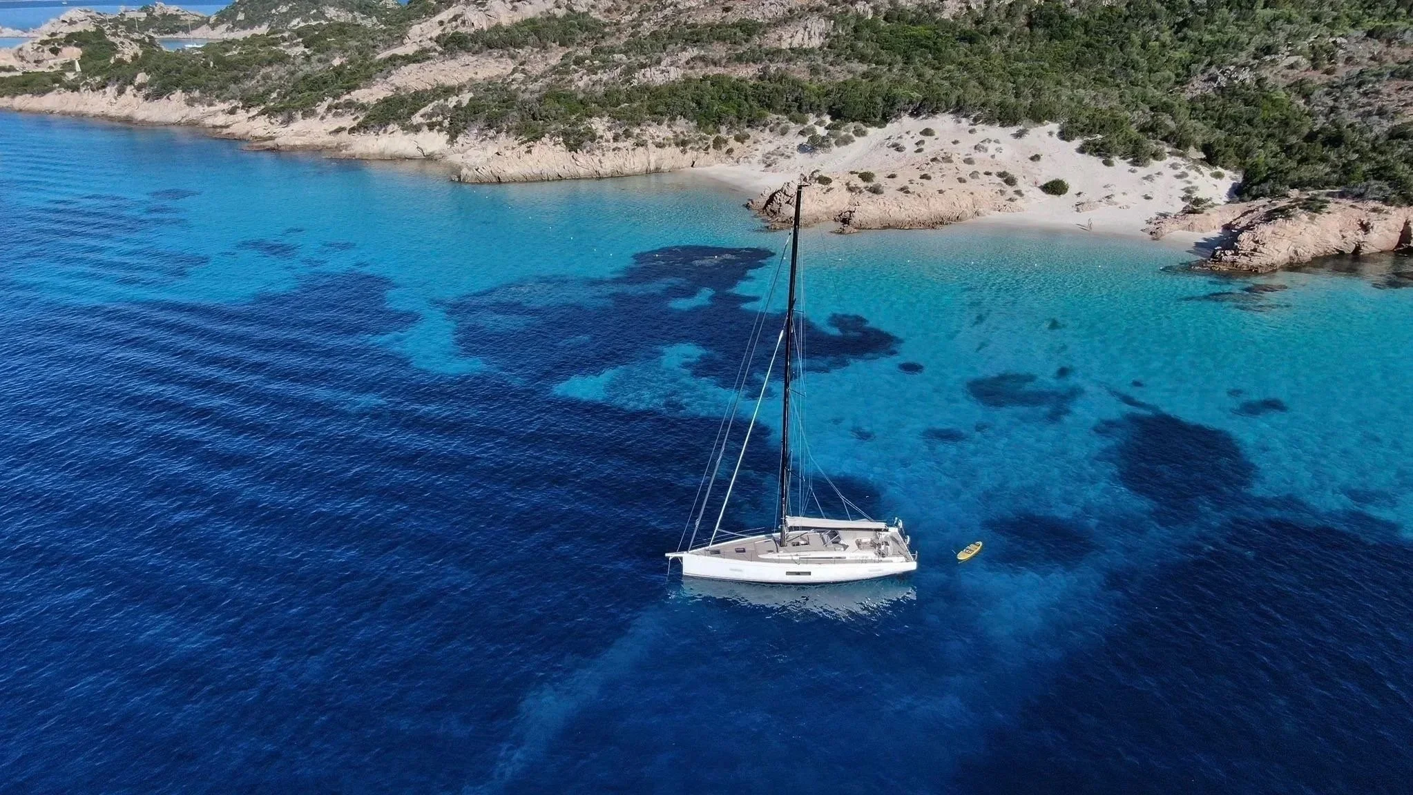 Aerial view of a first 53anchored in turquoise waters beside a rocky Mediterranean cove and sandy beach