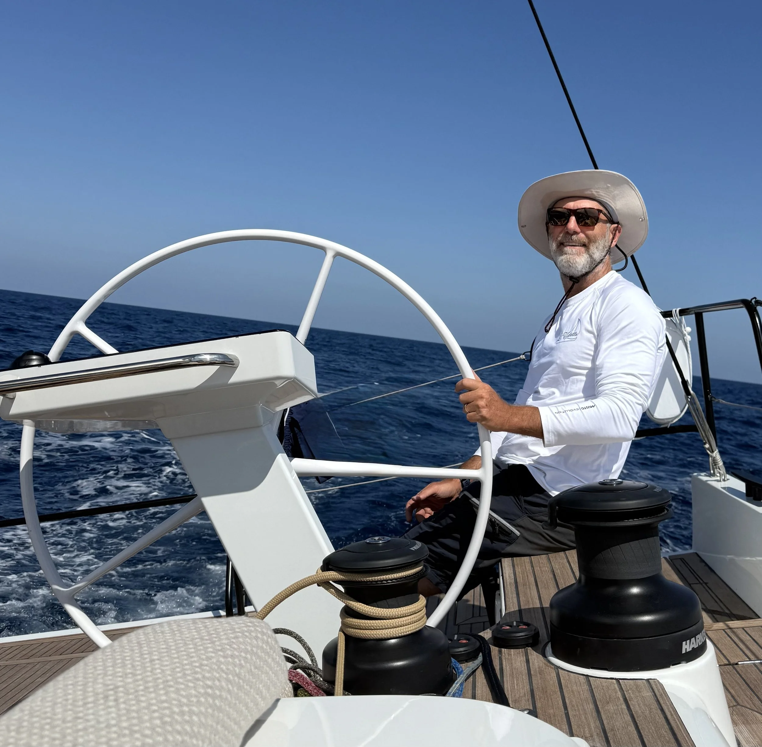 Matteo Our Captain smiling while sailing on a boat in the ocean, wearing a white hat and shirt, with clear blue sky above.