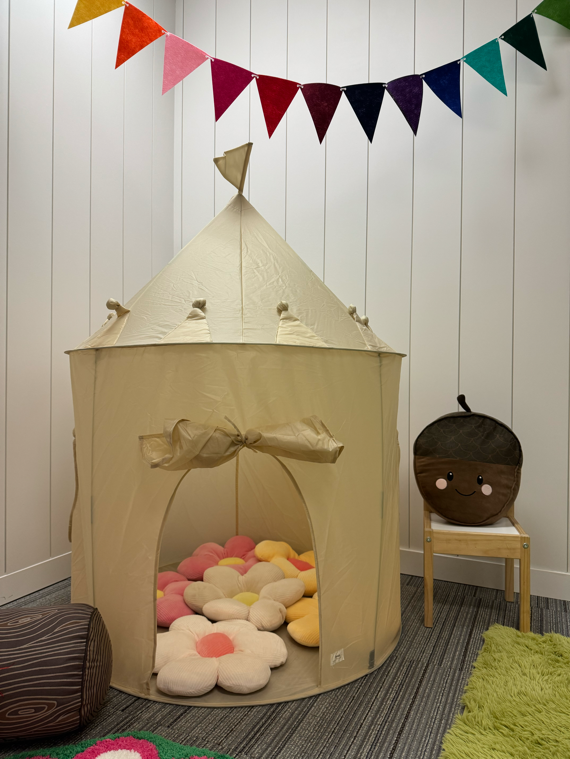 A children's play tent shaped like a castle with a yellow fabric and a bow at the entrance, filled with plush flower pillows. There is a brown plush acorn with a smiling face on a small wooden chair next to the tent. Colorful triangular pennant banne