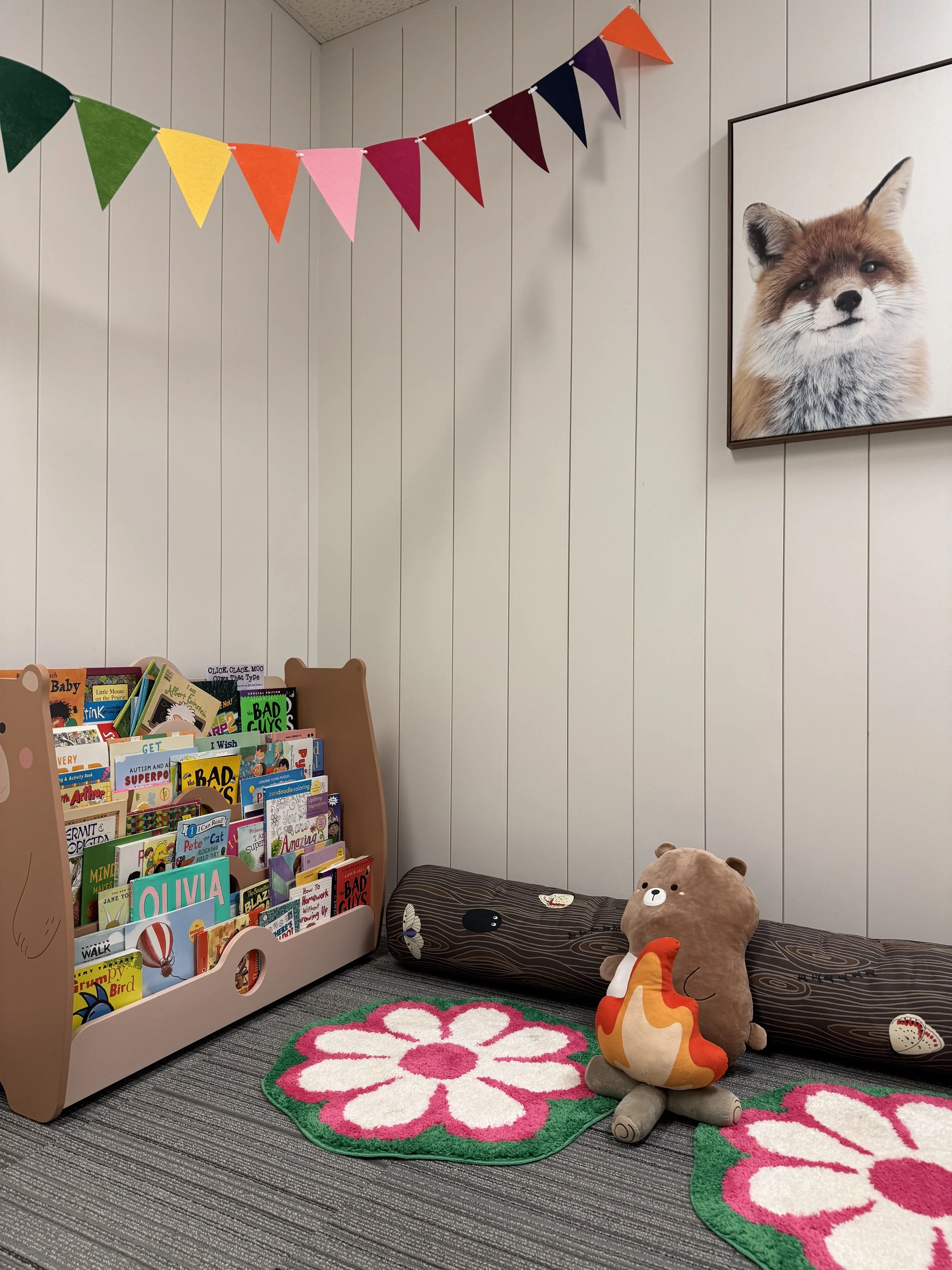 Children's reading nook with a beige book rack filled with colorful children's books, two pink and green flower-shaped rugs, a plush bear-shaped pillow with a fire pattern on its tummy, a large brown cylindrical pillow with baseball appliqués, a colo