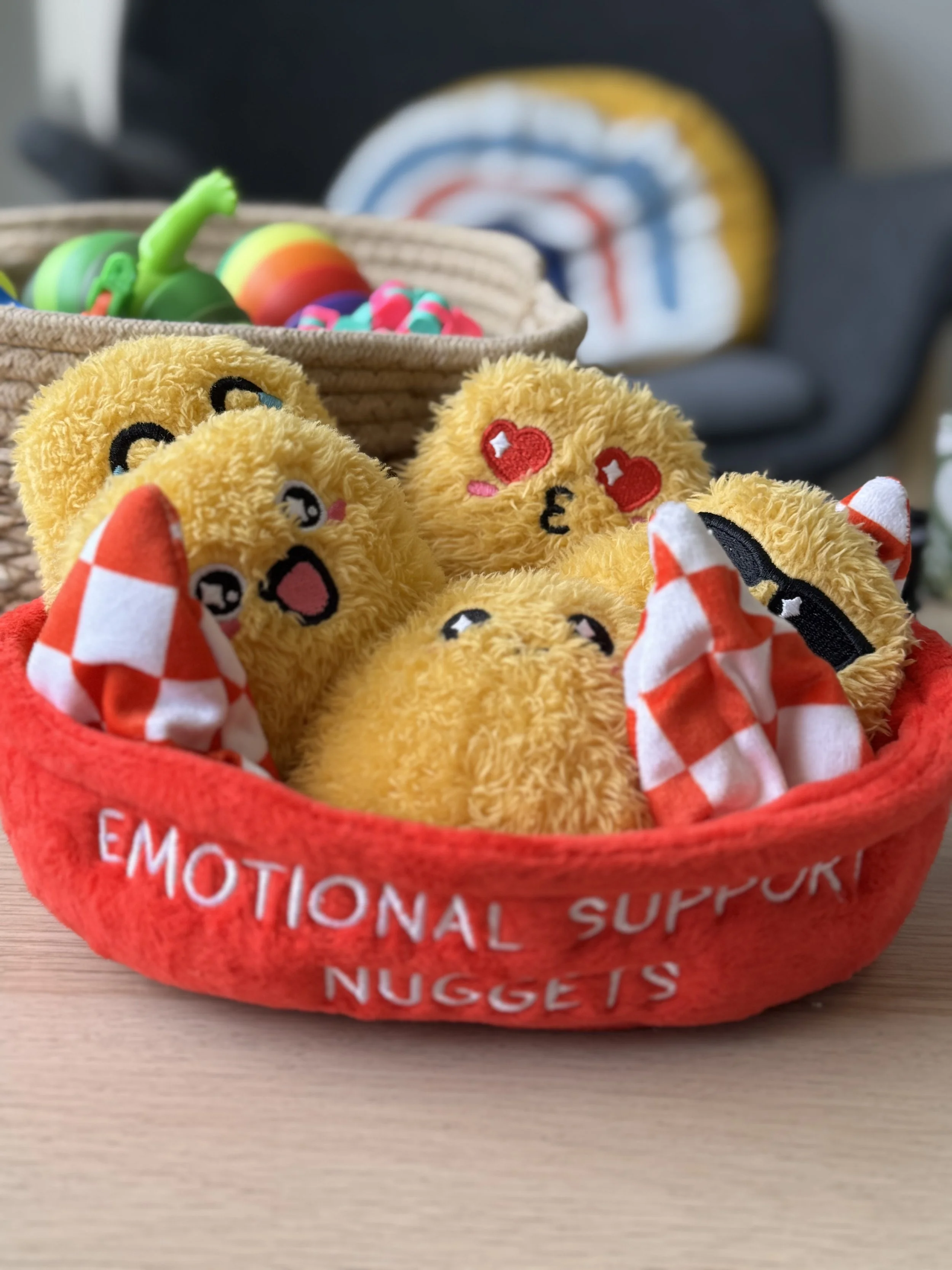 A basket of plush teddy bear toys with embroidered faces, inside a red fabric container labeled 'EMOTIONAL SUPPORT NUGGETS,' with a basket of colorful rubber toys in the background.