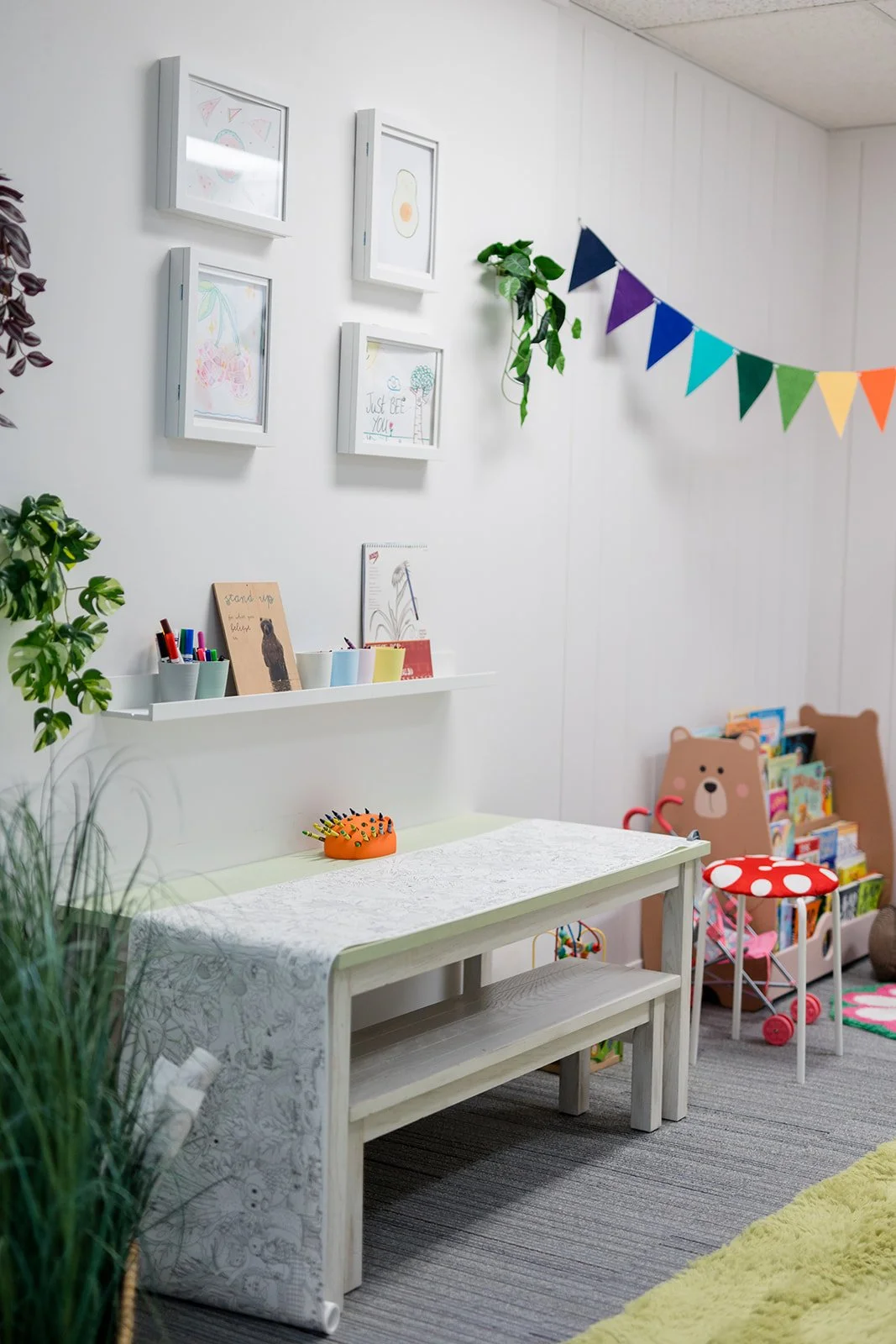 Children's playroom with a white table, colorful toys, picture frames on the wall, a rainbow bunting, and bookshelves with children's books and stuffed animals.