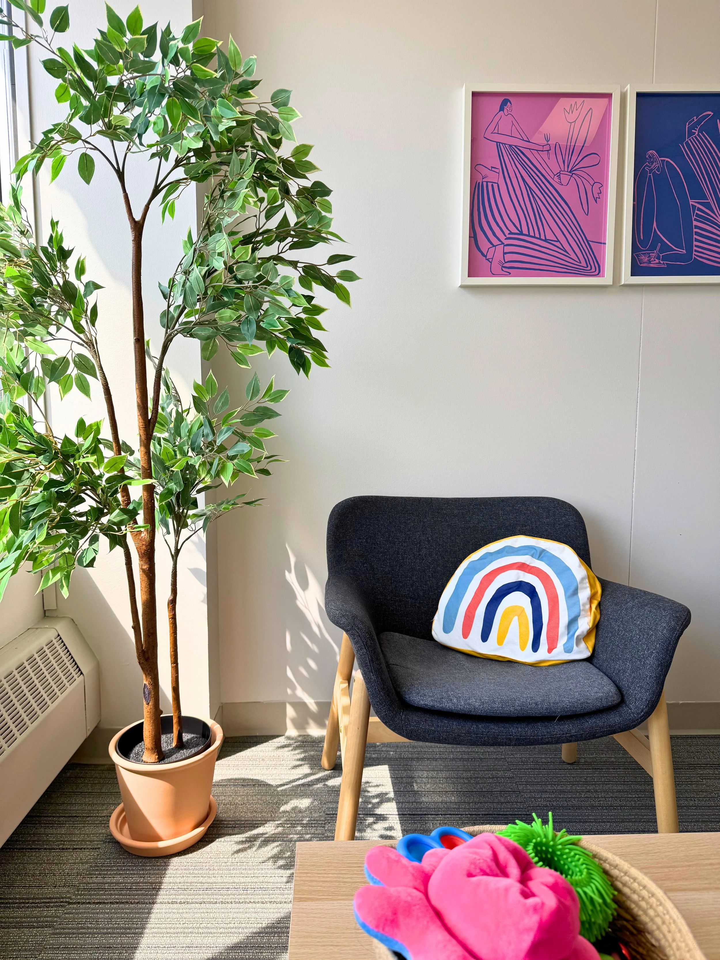A cozy seating area featuring a dark gray armchair with a rainbow-patterned pillow, a tall artificial plant in a terracotta pot beside it, and two framed abstract line art prints on the wall in pink and blue colors.