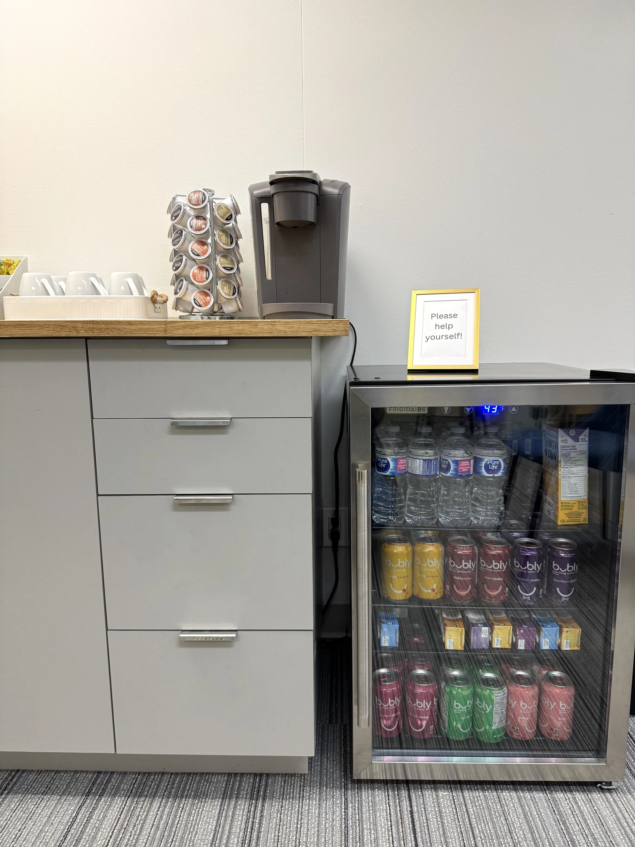 A small refreshment station with a coffee maker, a container with coffee pods, a white tip jar, and a fridge filled with bottled water and canned beverages. A sign on the fridge reads 'Please help yourself!'