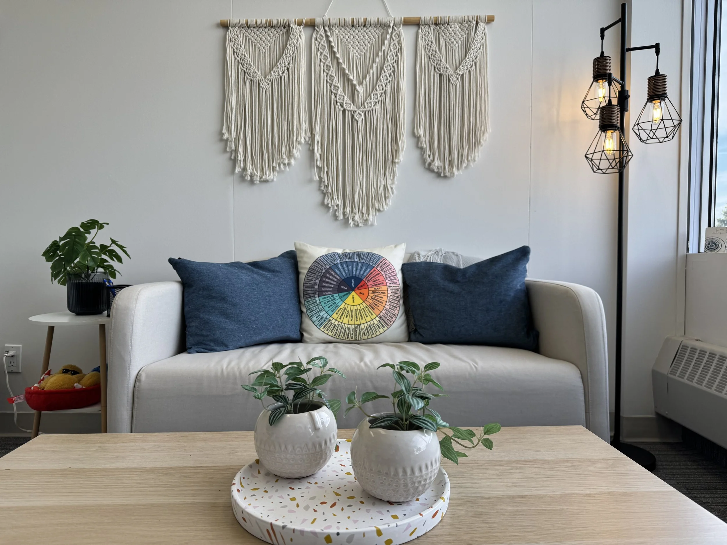 Living room with a white sofa decorated with three navy cushions and a colorful wheel pillow, a wooden coffee table with two potted plants, a green plant on a small side table, macrame wall hanging above the sofa, and a black floor lamp with three ge