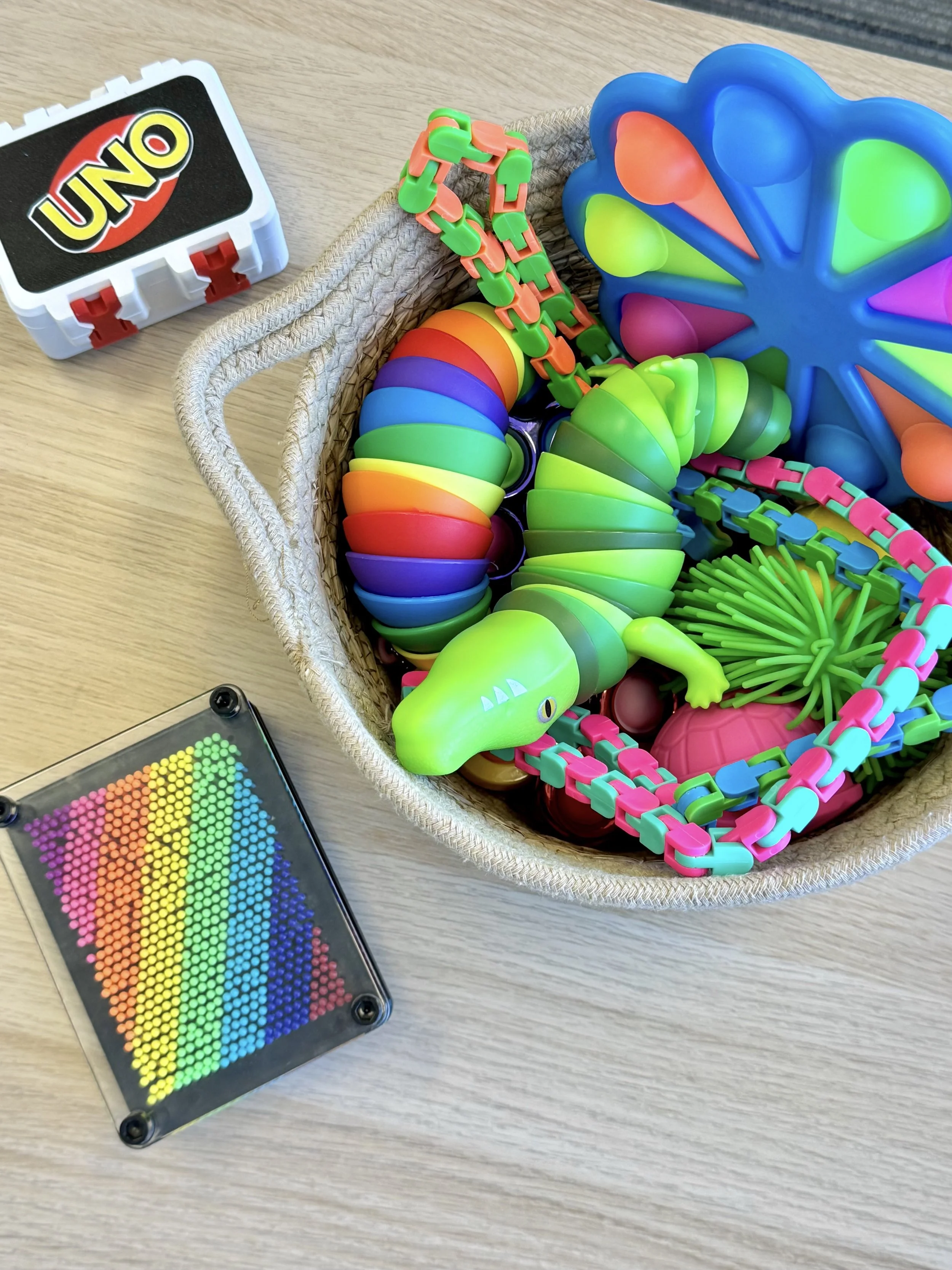Colorful toys in a basket, including a rainbow-colored caterpillar, a green dinosaur, a pink ball, a spiky green ball, and chain links, with a rainbow-colored dot grid and an UNO toy on a wooden surface.