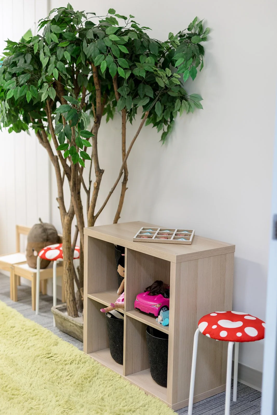 Indoor play area with a tall leafy green tree, a light wood shelf with toys, a small bench with mushroom-shaped cushion, and a yellow shaggy rug.