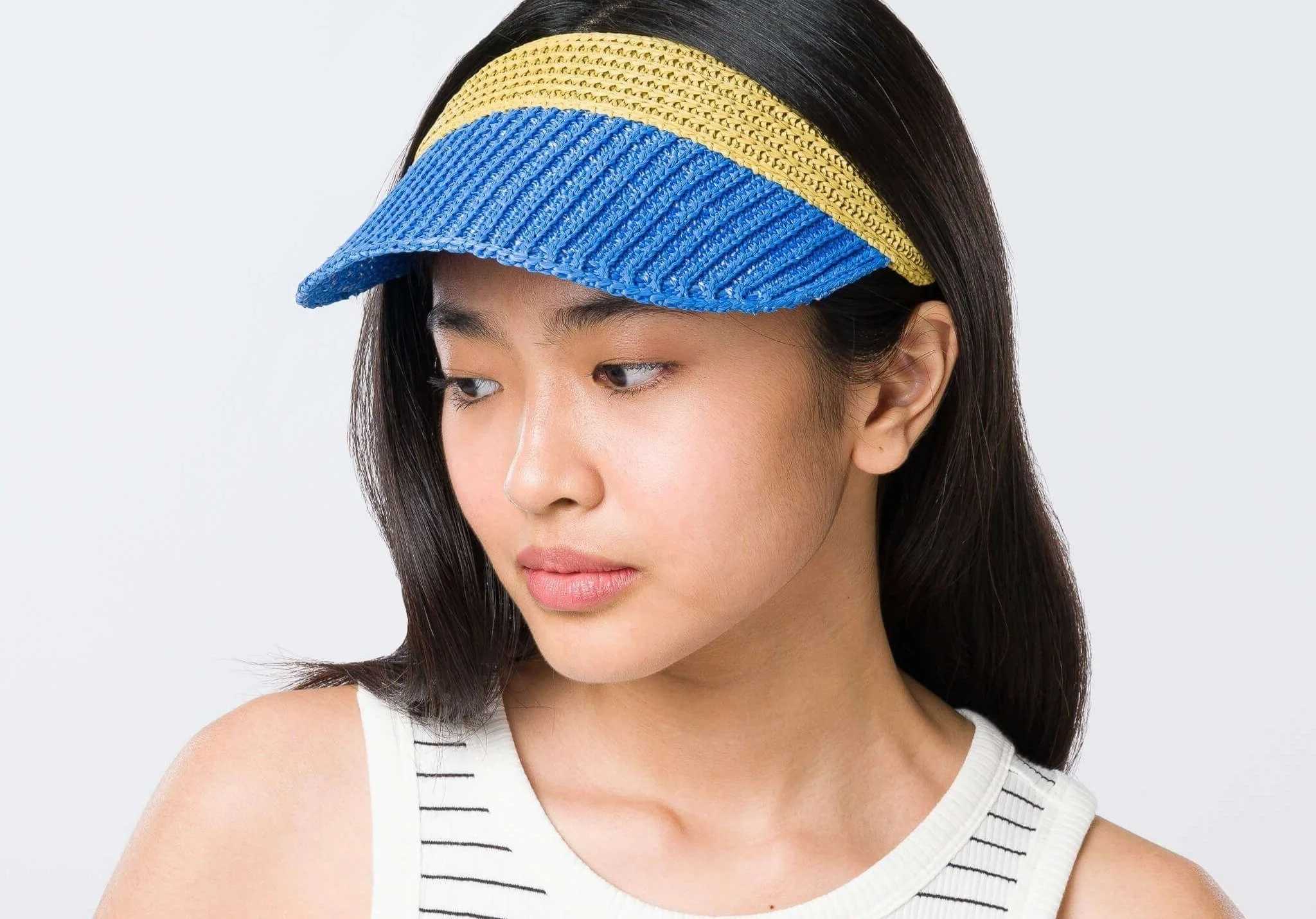Young woman with long dark hair wearing a multicolored handmade crochet, woven, raffia visor, looking to her left against a plain light background.
