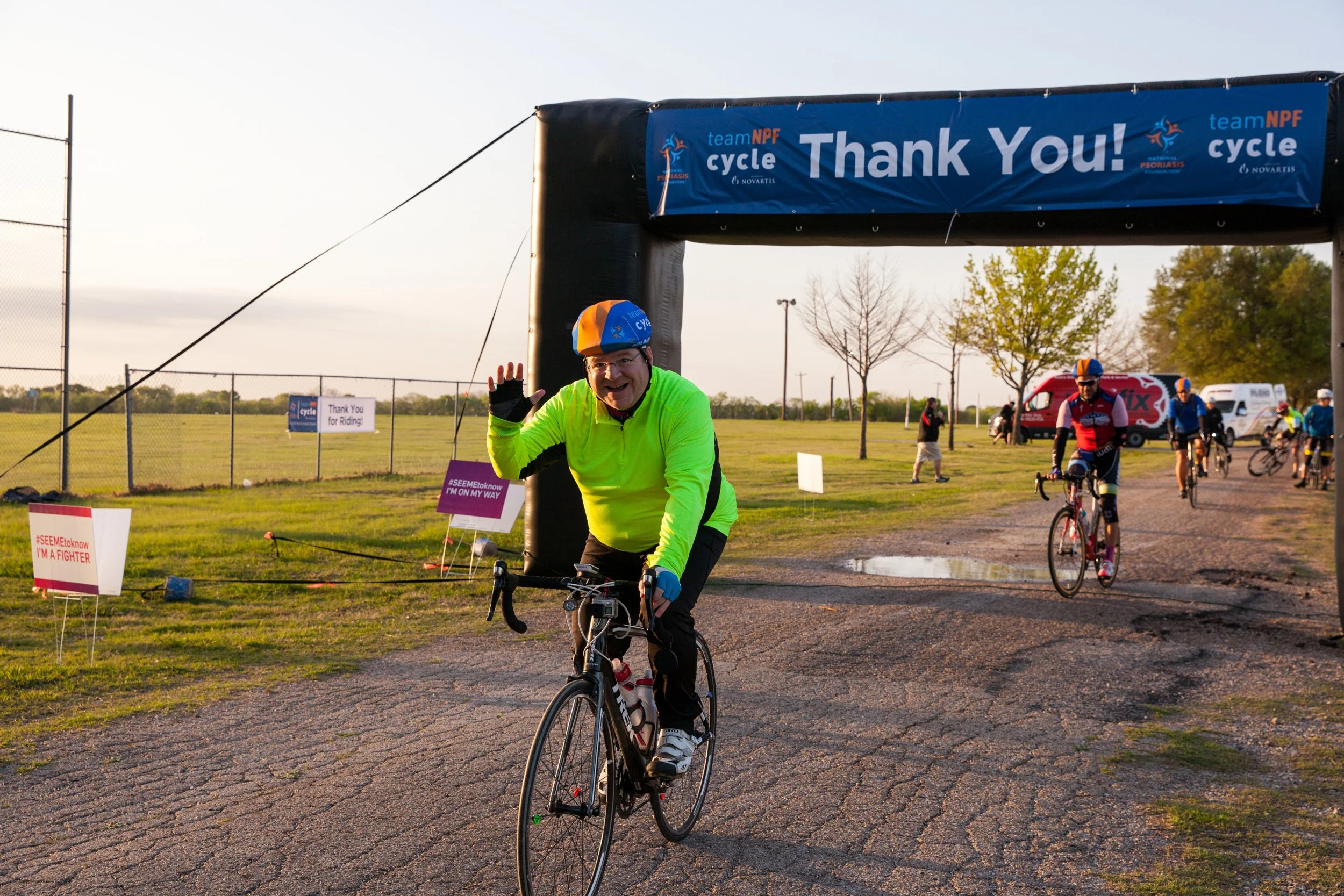 A cyclist smiles and waves while riding through the Team NPF event arch during a nonprofit fundraising ride in the Dallas-Fort Worth area.