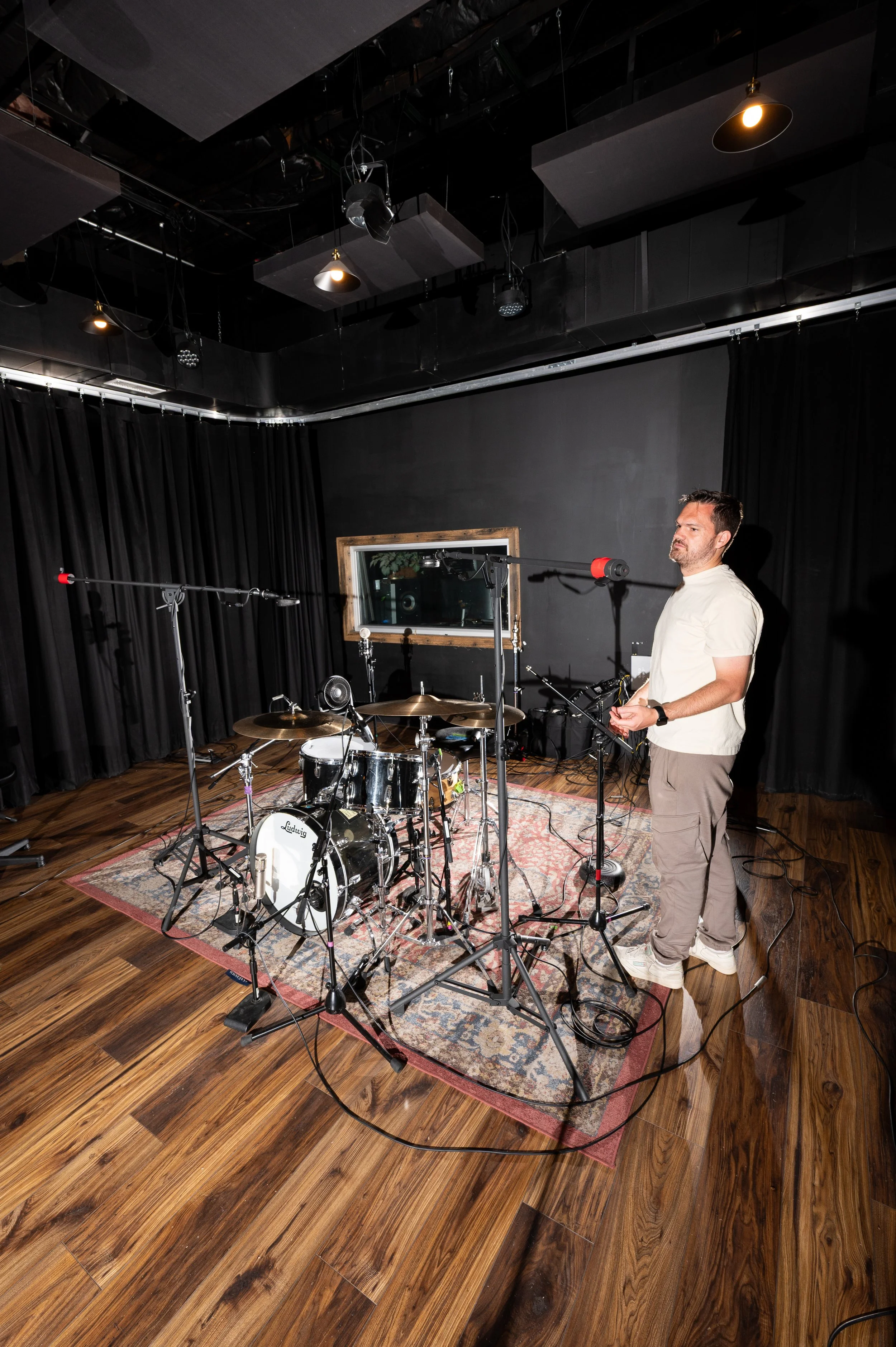 A smiling young man sitting in a recording studio workspace surrounded by audio equipment, with a computer monitor displaying audio editing software in the background.