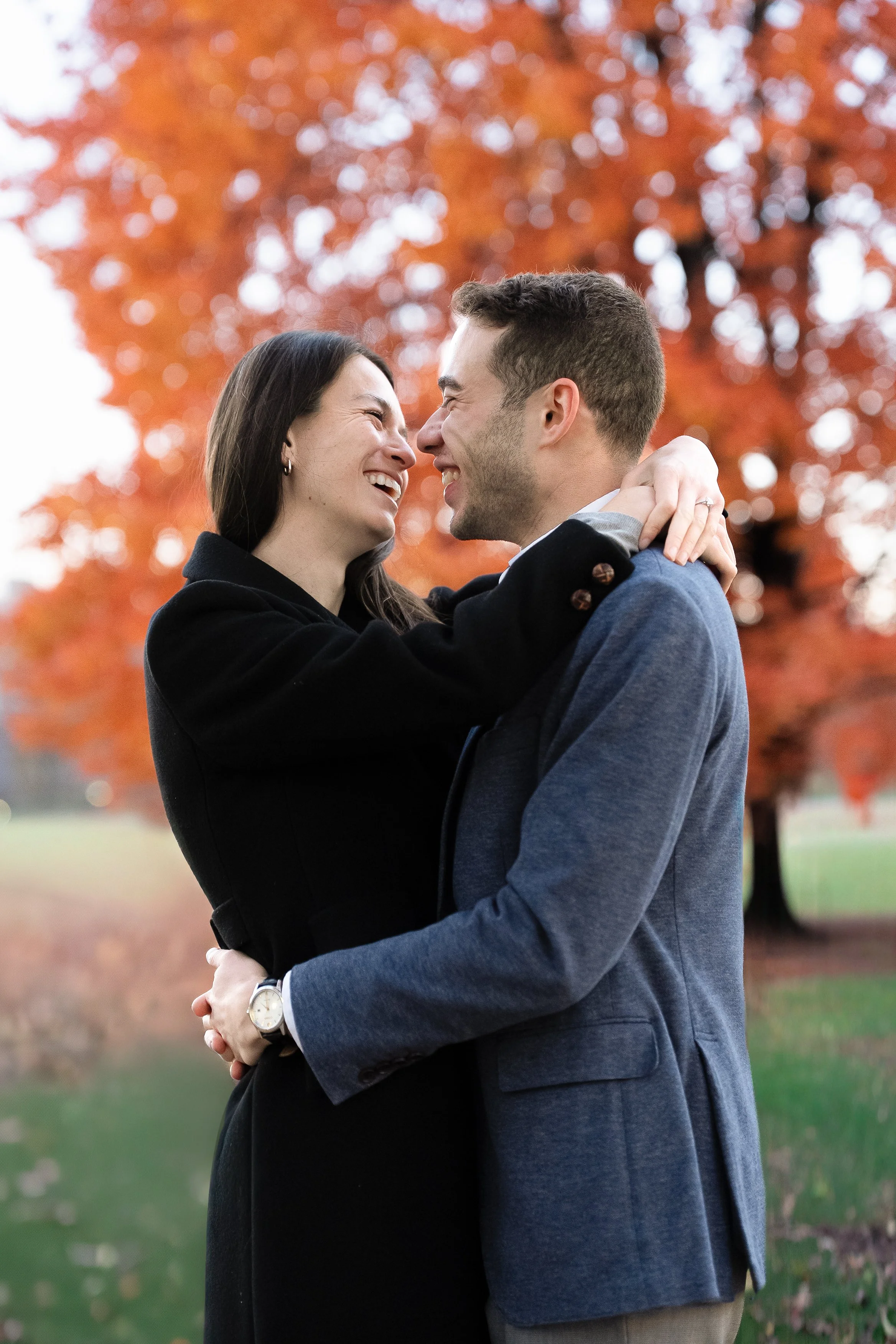 A couple is embracing outdoors during autumn, with a background of orange leaves on trees. They are smiling and holding each other closely.