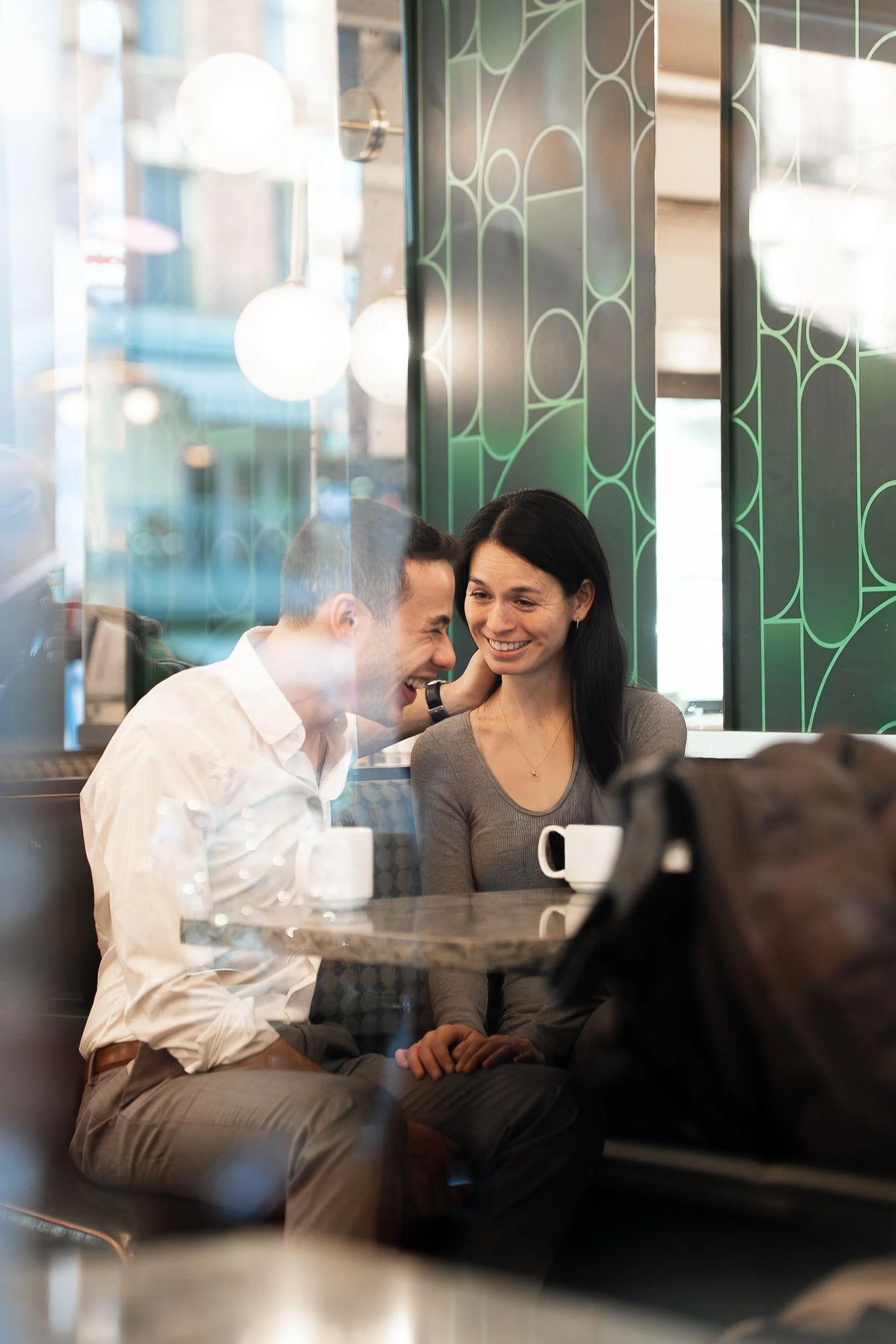 A man and woman sitting at a table in a cafe, smiling and appearing to share a joyful moment, with coffee cups on the table and a reflective glass window in front of them.