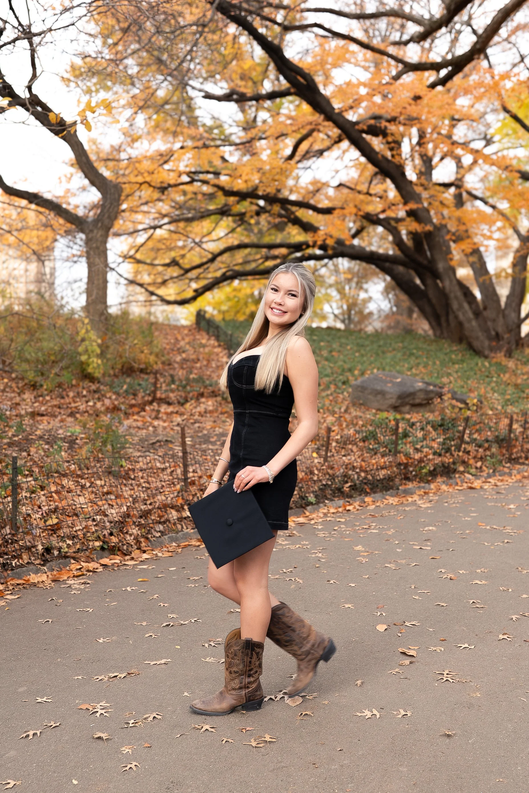 A young woman in a black dress and cowboy boots holding a graduation cap, smiling outdoors during autumn with fallen leaves and orange trees in the background.
